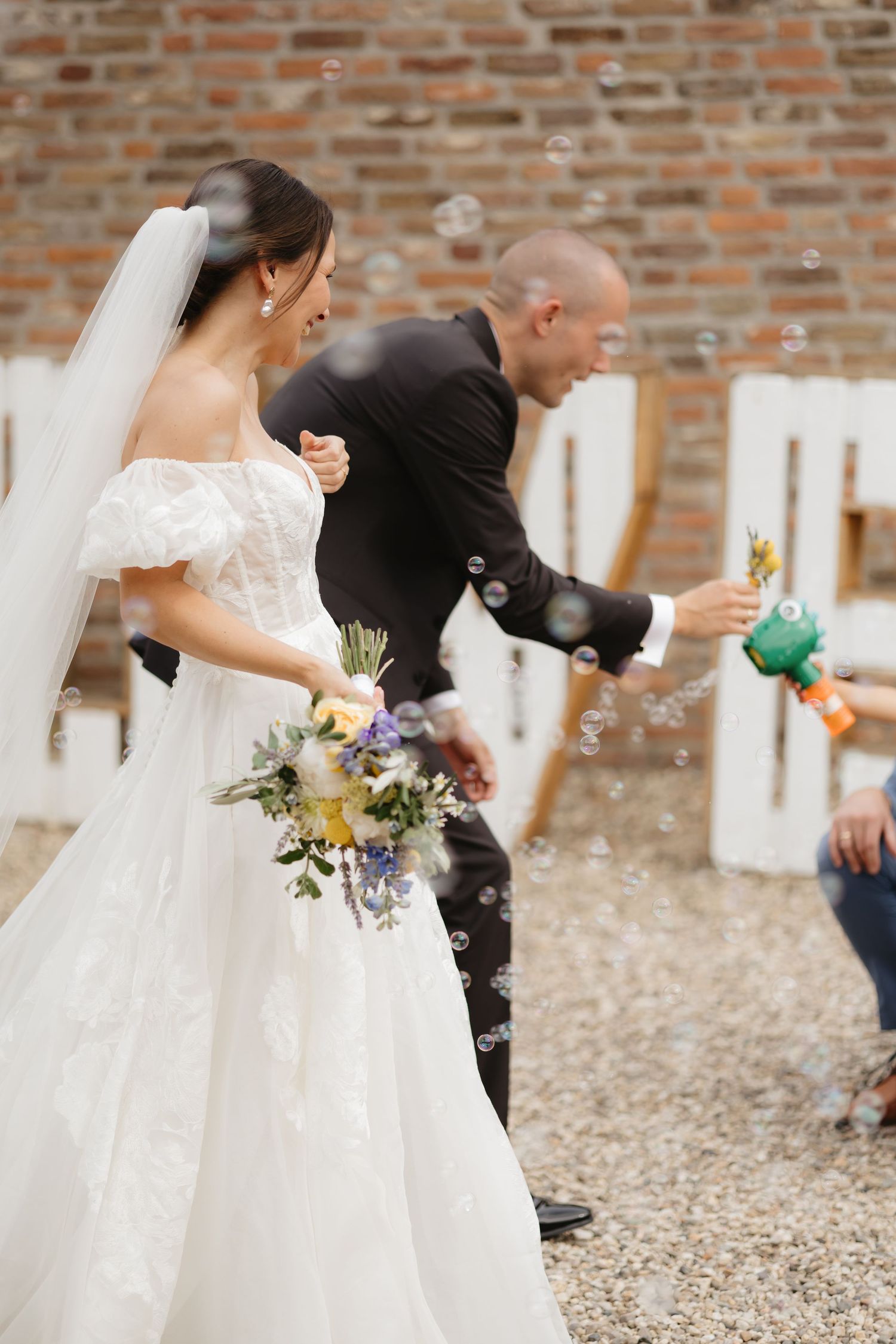 Wedding celebration on cobblestone ground with guests blowing bubbles near brick wall.