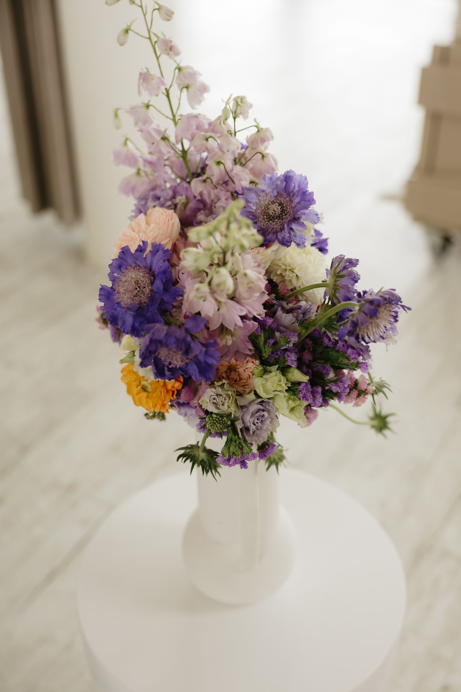 A delicate floral arrangement with purple, pink, and yellow blooms in a white vase on a light surface.