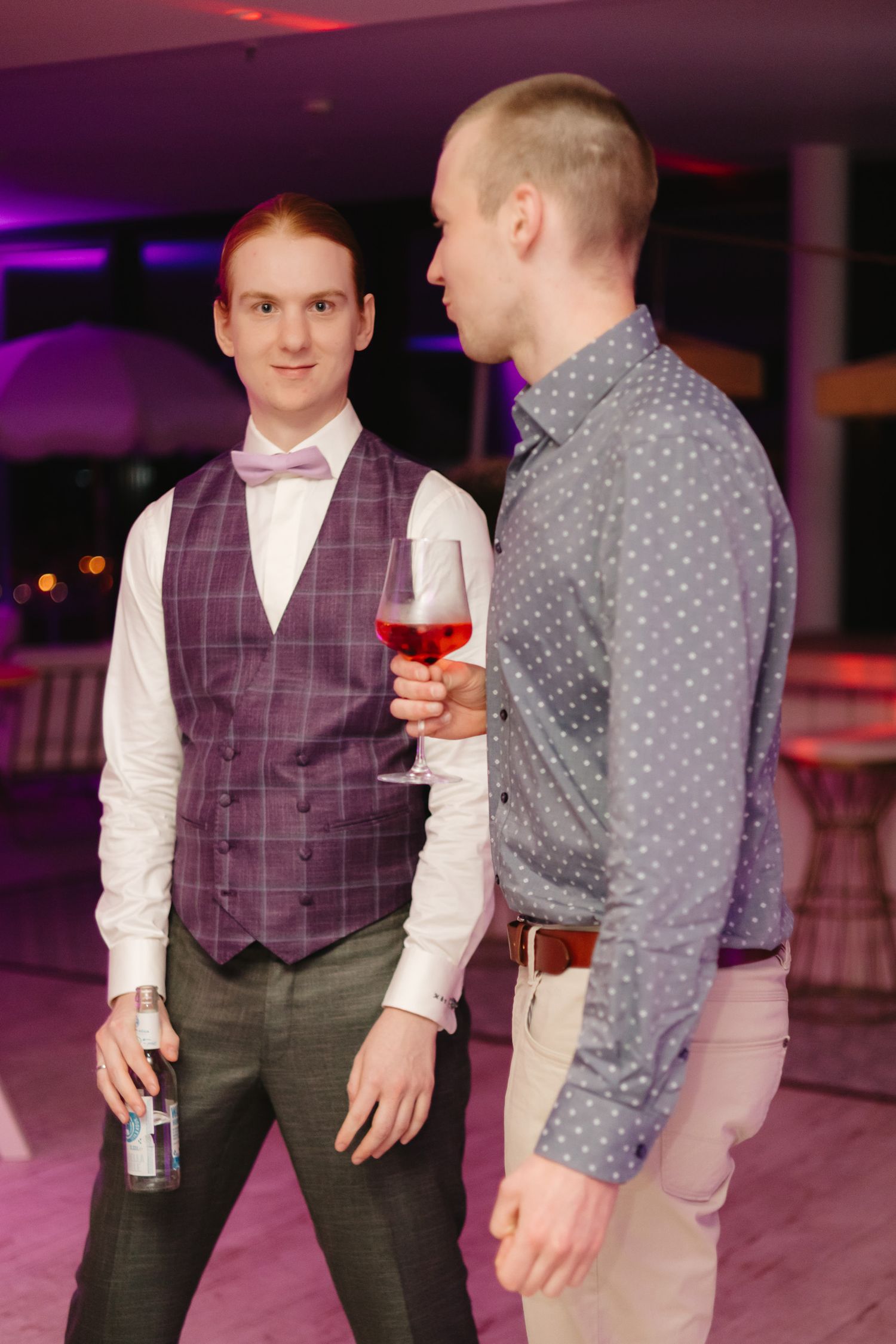 Two people in formal attire having drinks and conversation in a venue with purple mood lighting.