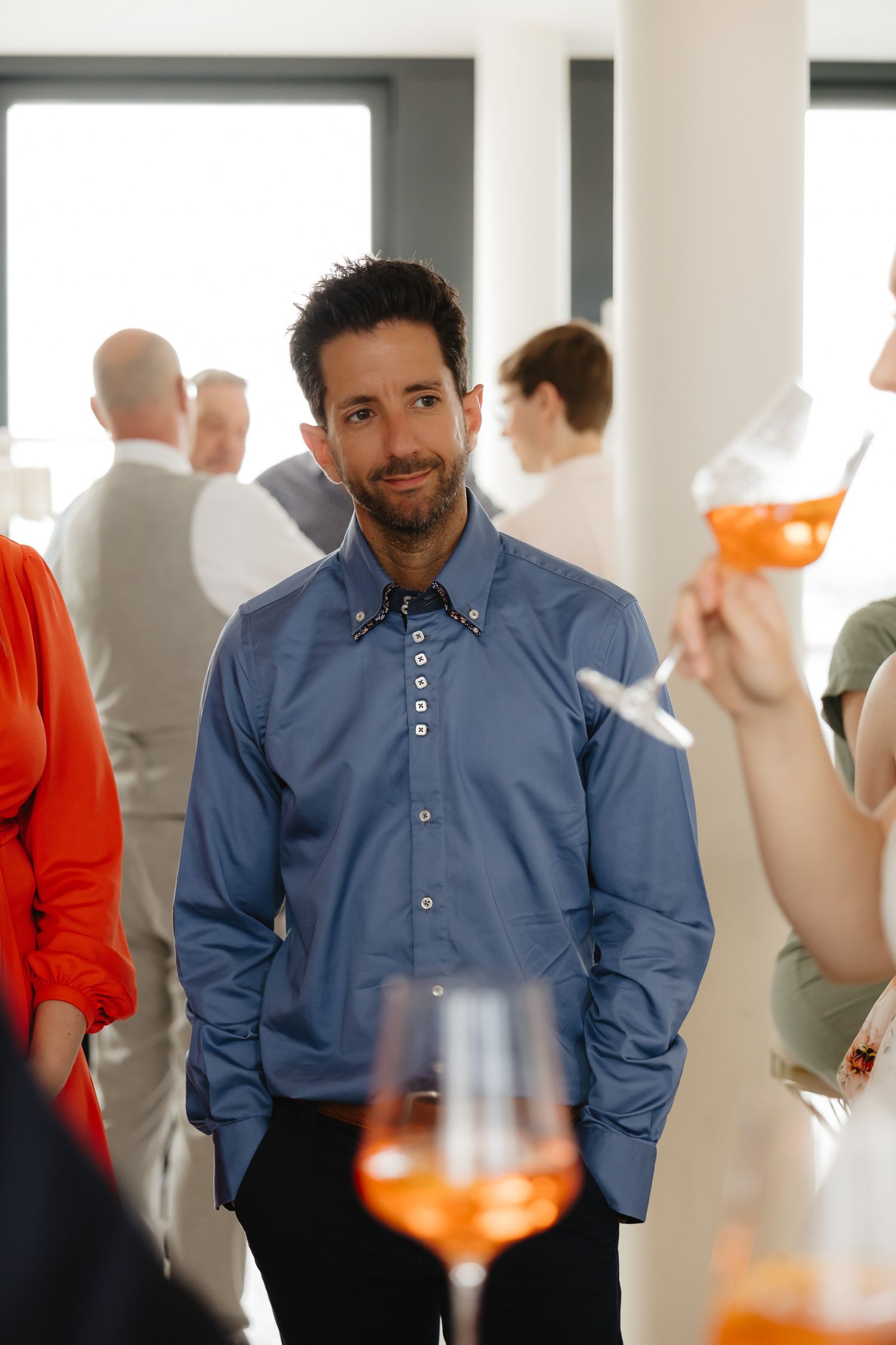 A person in a blue button-down shirt stands at a social gathering with drinks and other attendees in the background.