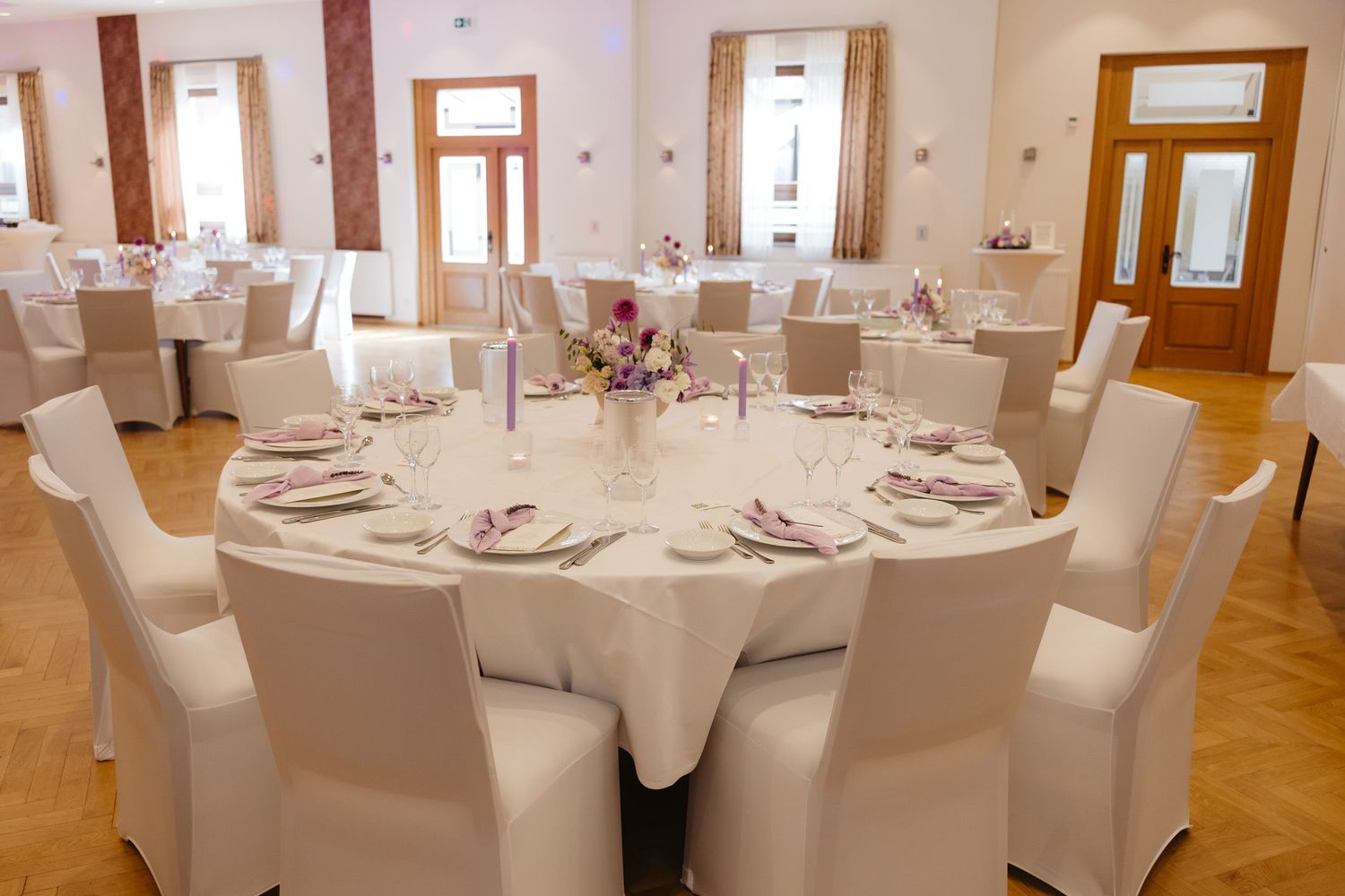 A bright banquet hall with round tables covered in white linens and elegant place settings arranged in a formal dining setup.