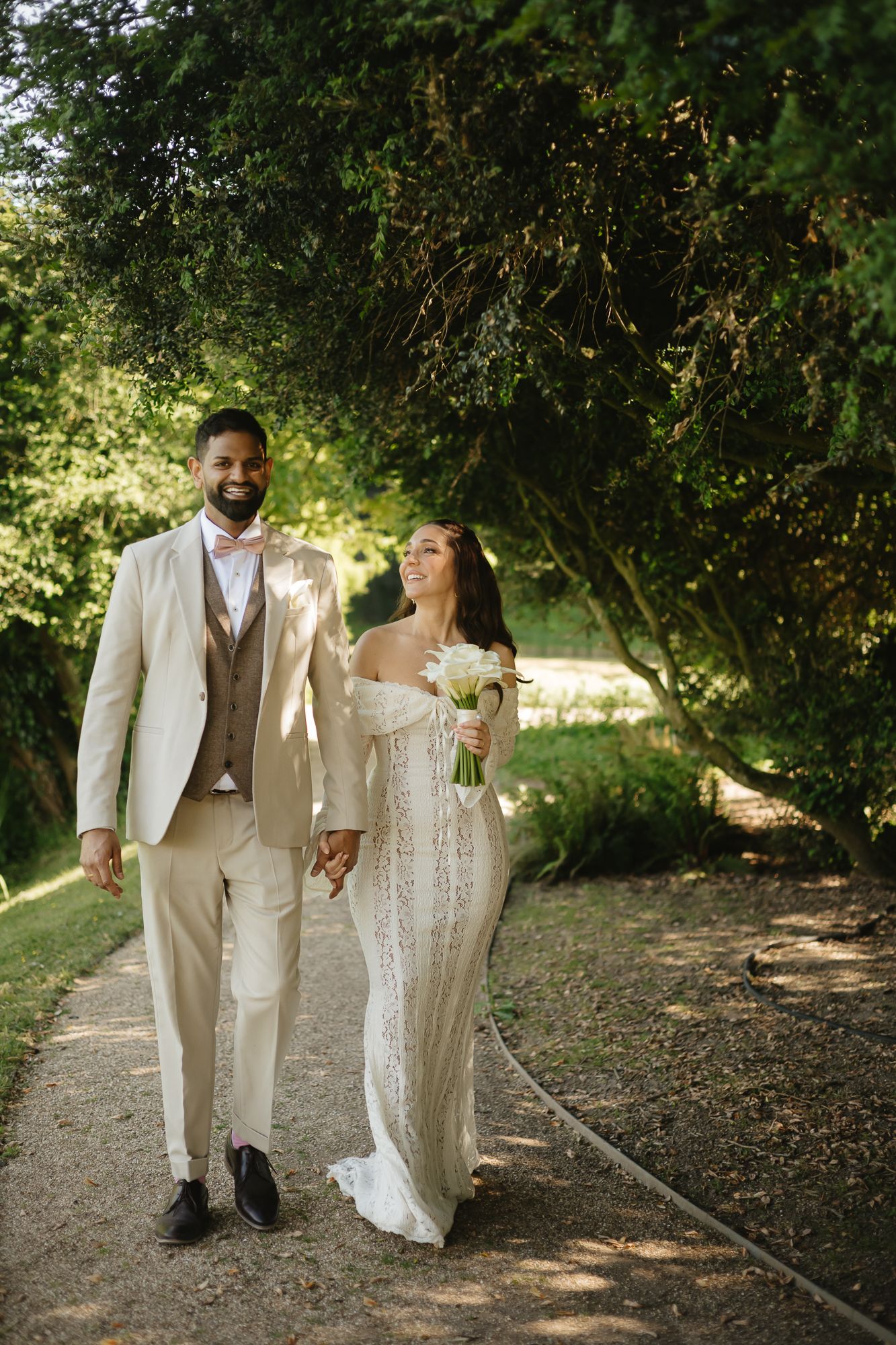 Couple in cream and white wedding attire walking hand in hand along a tree-lined path.