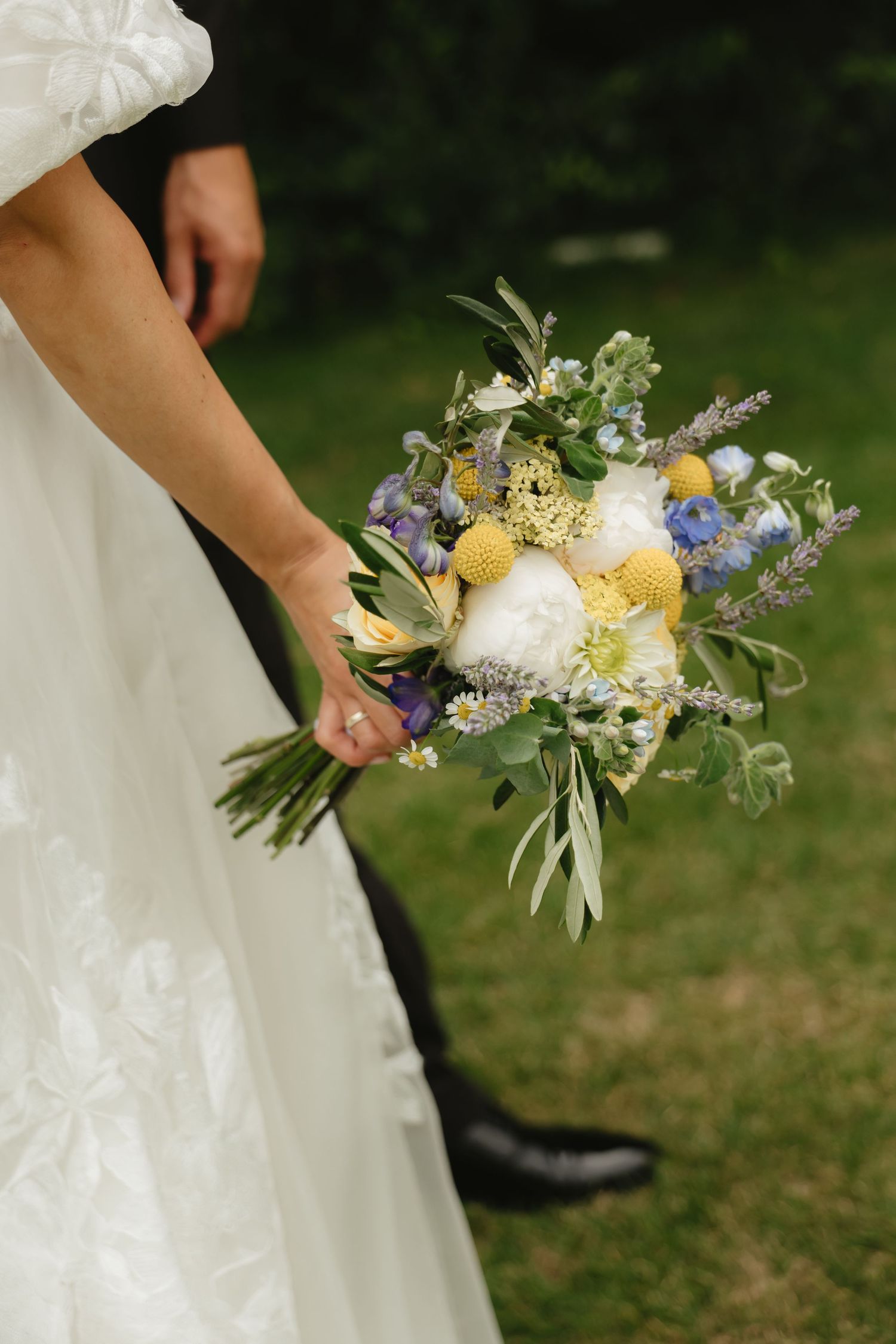 Close-up of bridal bouquet featuring yellow, white, and blue flowers with delicate greenery.