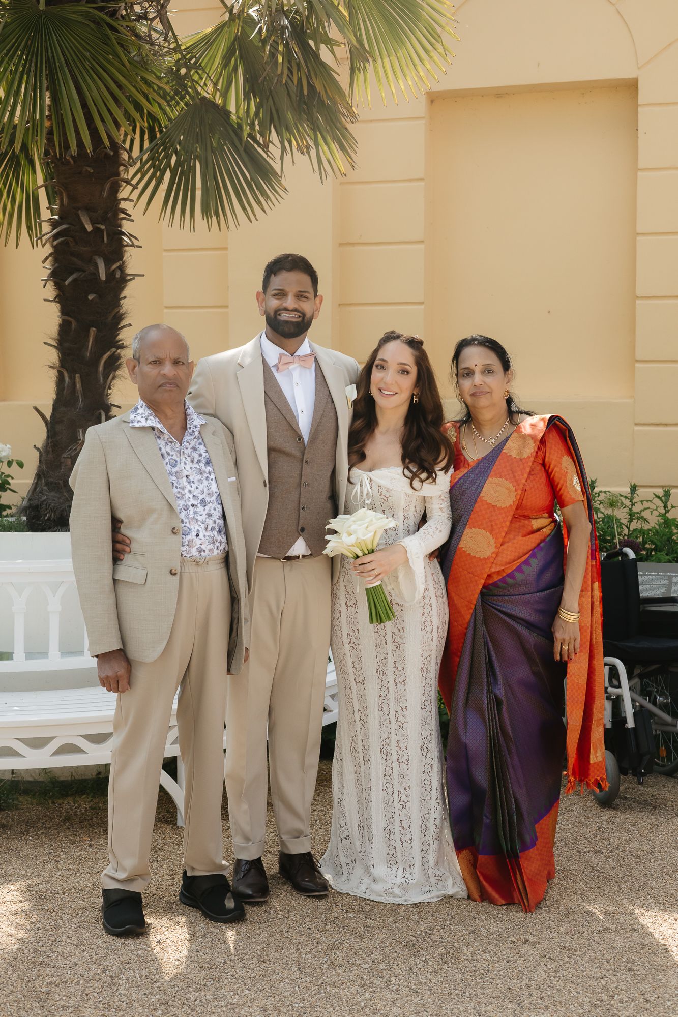 Wedding party poses for a group photo outdoors with palm trees and beige building in background.