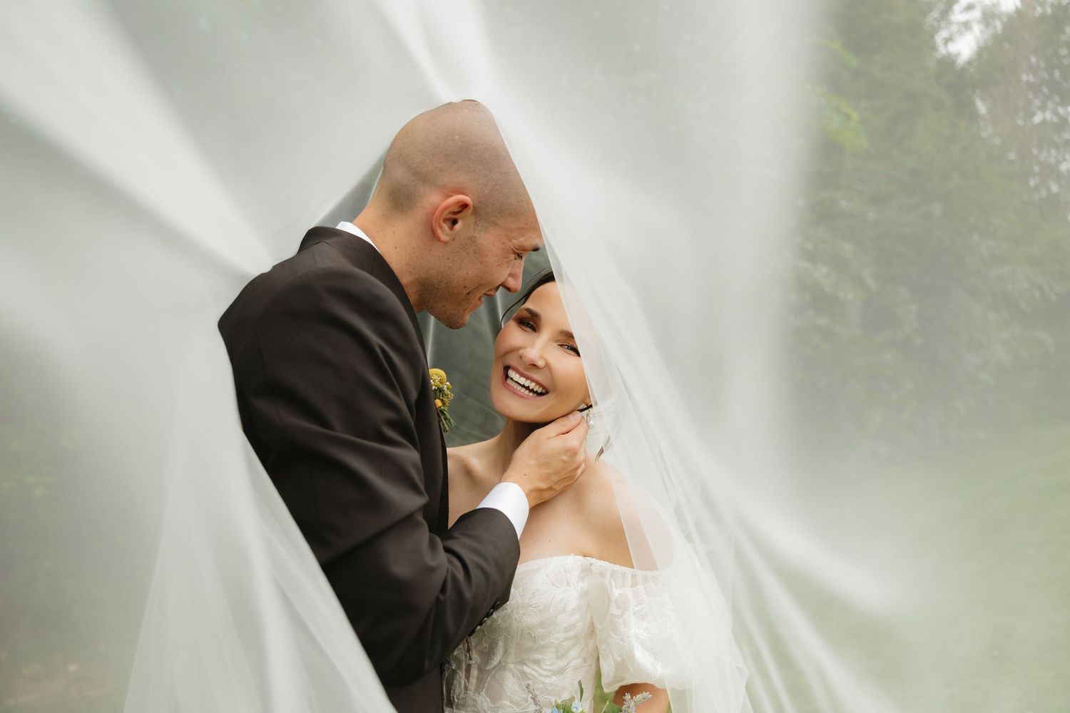 Newlyweds share a tender kiss beneath a delicate white veil in an outdoor setting.