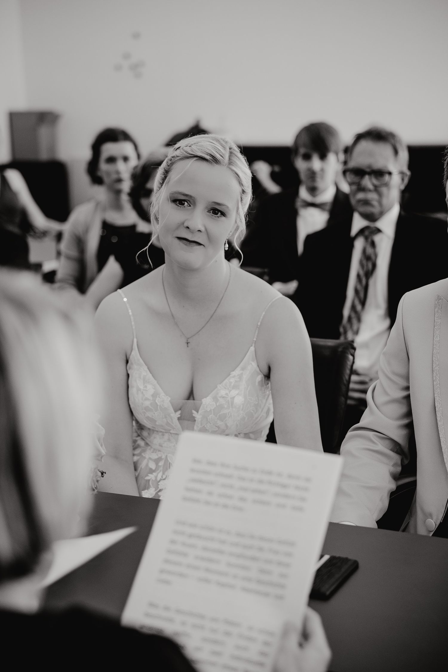 Black and white photo showing a bride in a white dress during a wedding ceremony while guests look on.