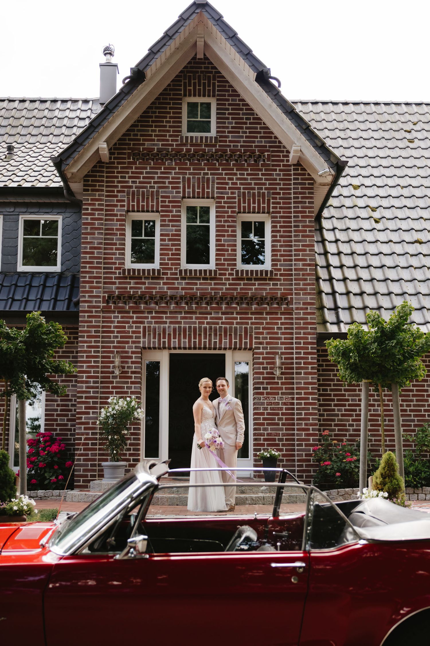 A vintage red convertible car parked in front of a traditional brick house with white trim and tiled roof.