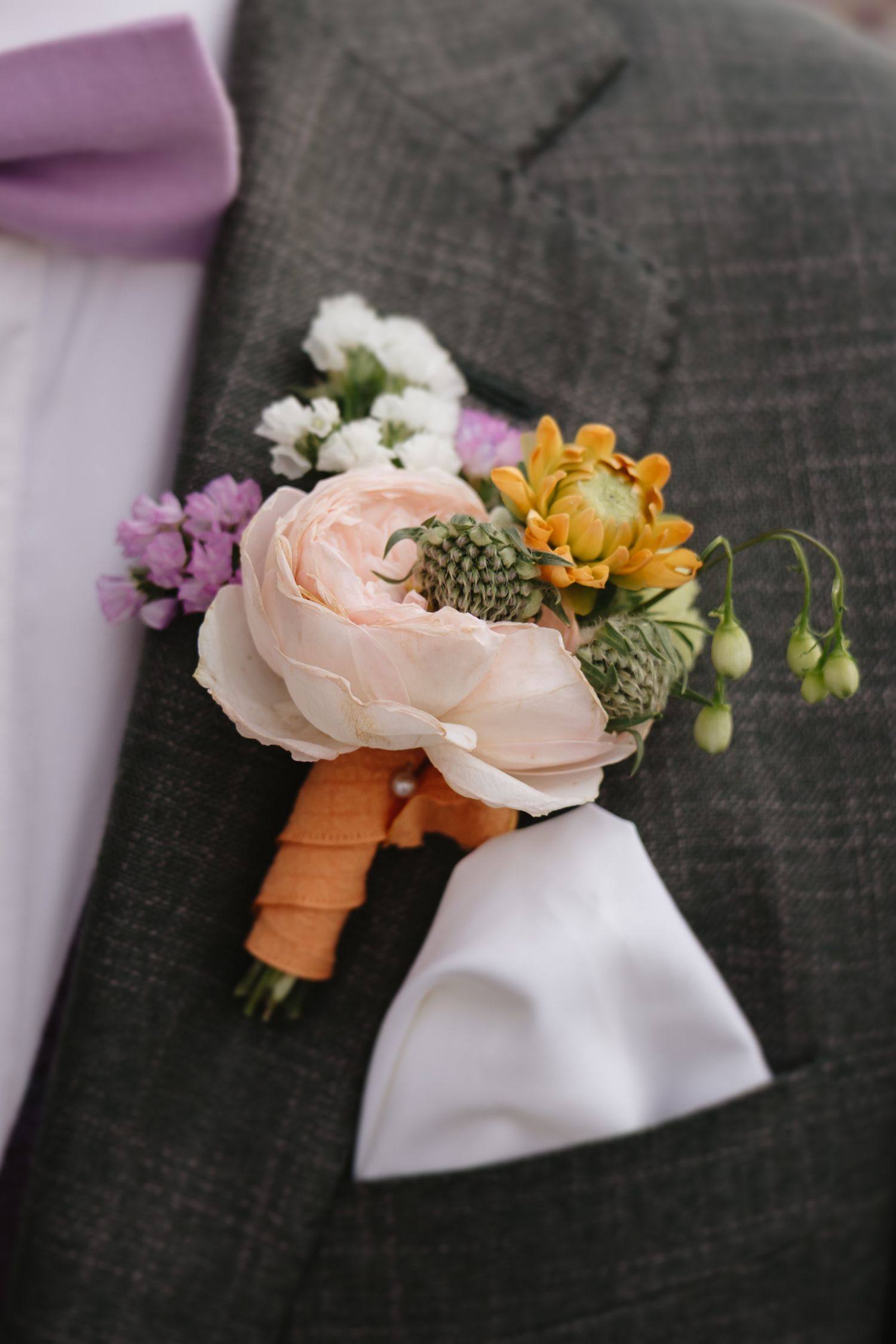 A delicate boutonniere featuring pink roses, yellow flowers and greenery pinned to a dark gray suit lapel.