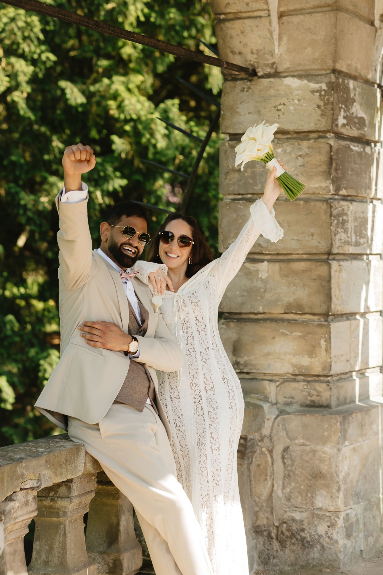 Celebratory wedding poses with couple holding bouquet under stone archway.