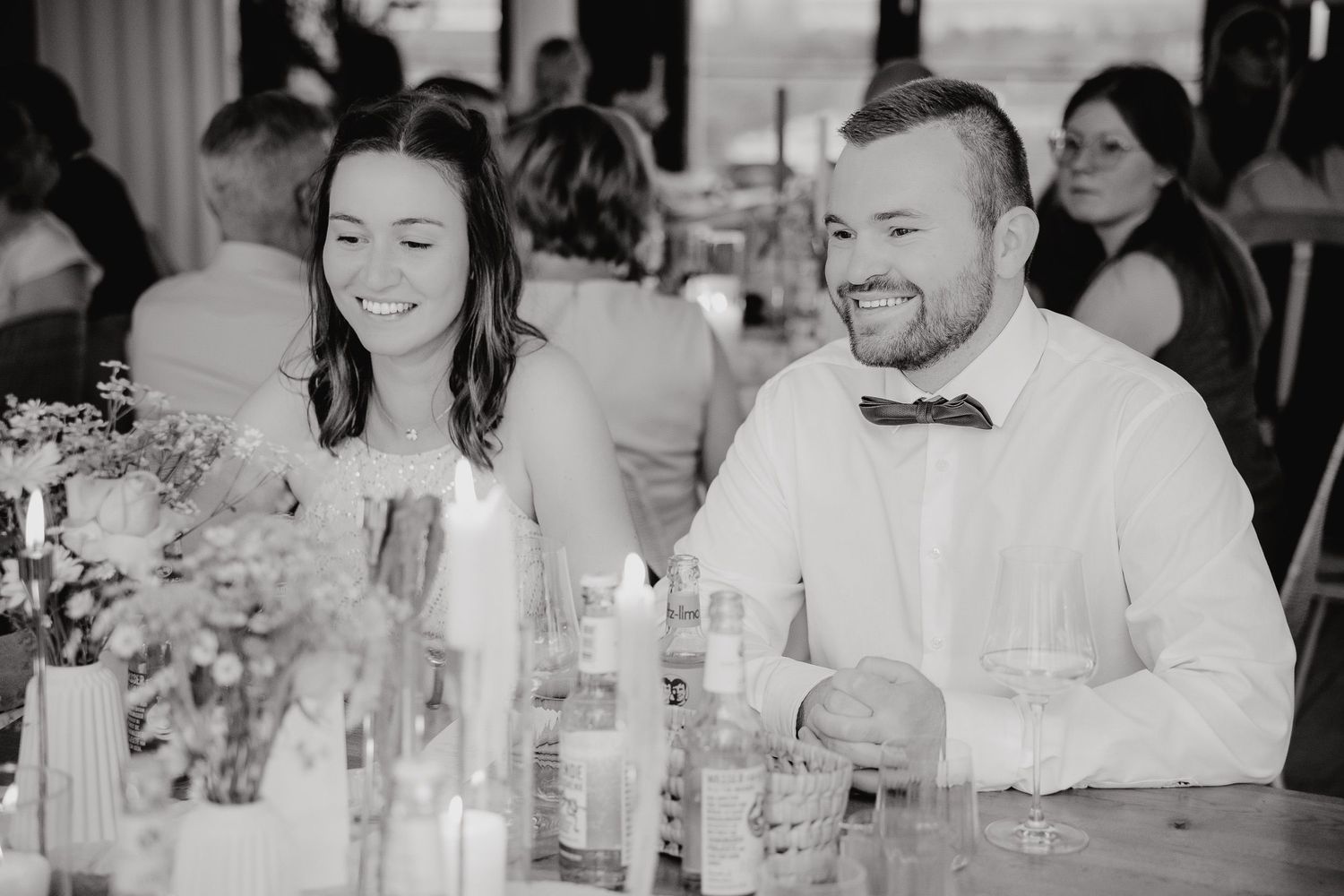 A black and white candid photo of a couple laughing together at a wedding reception table with floral centerpieces.