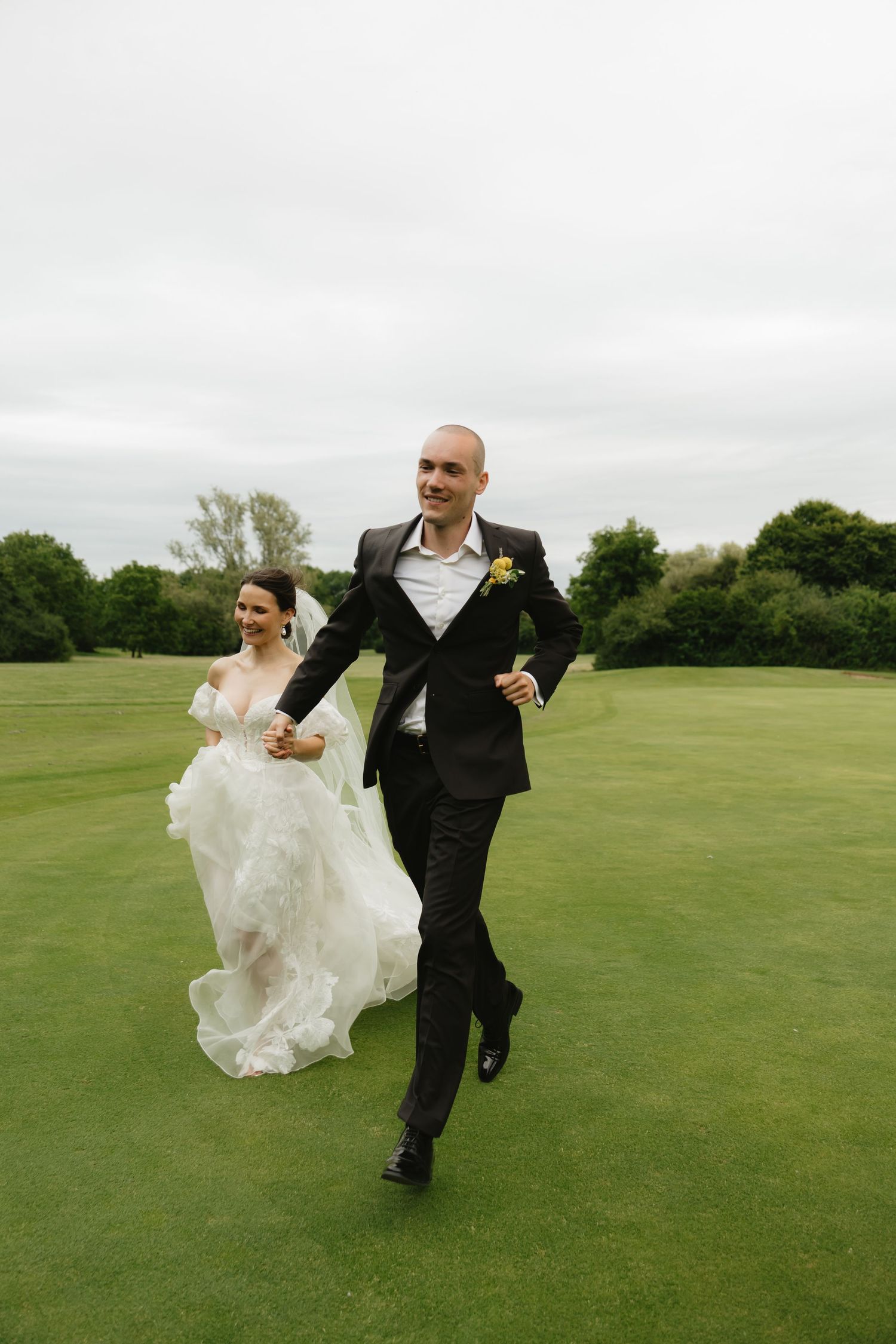 Newlyweds run playfully across a green golf course under cloudy skies.