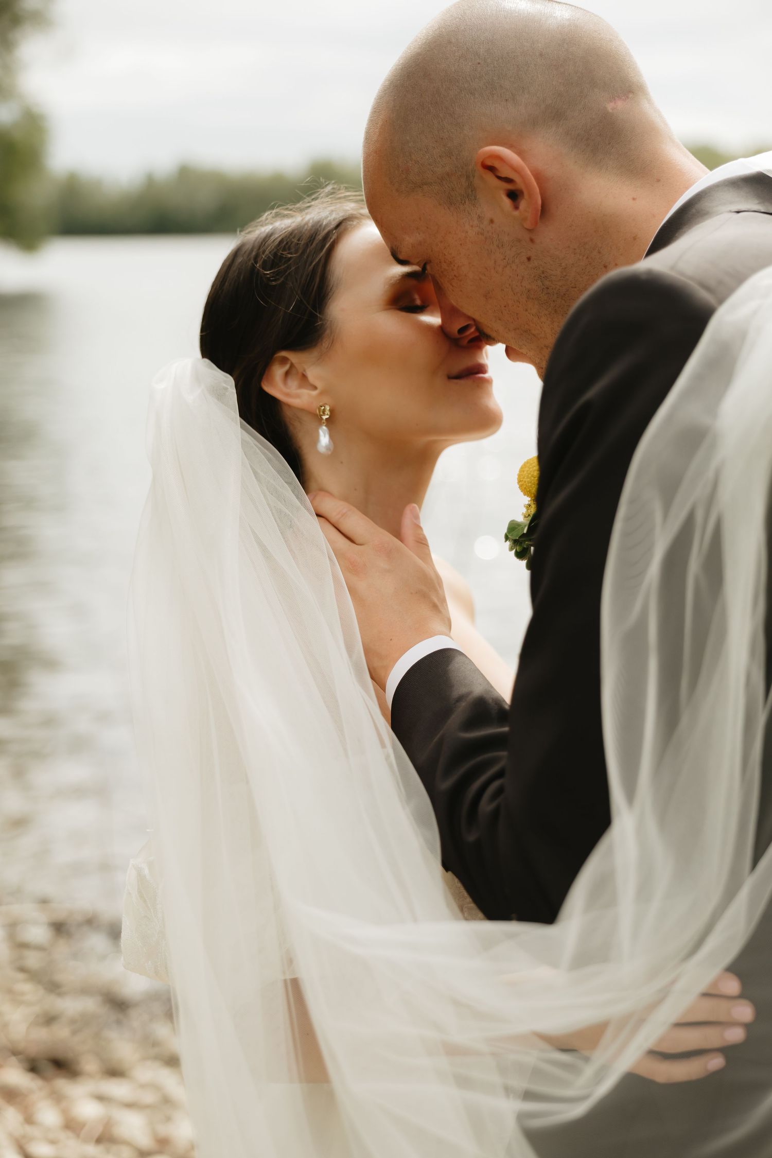 Romantic wedding couple embraces by the water as the bride's veil flows in the breeze.