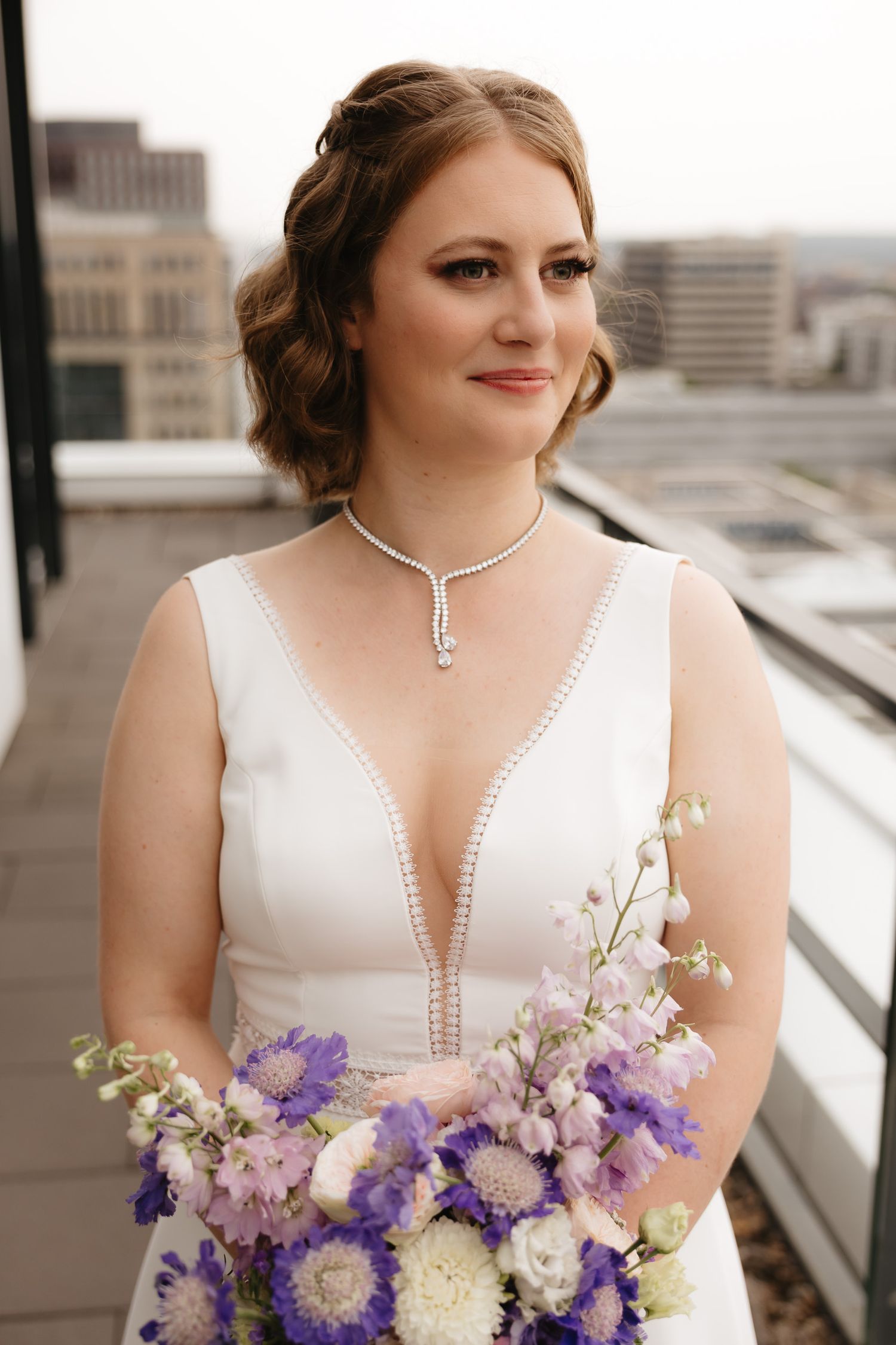 A bride in a white wedding dress holds a stunning purple and white floral bouquet on a rooftop setting.