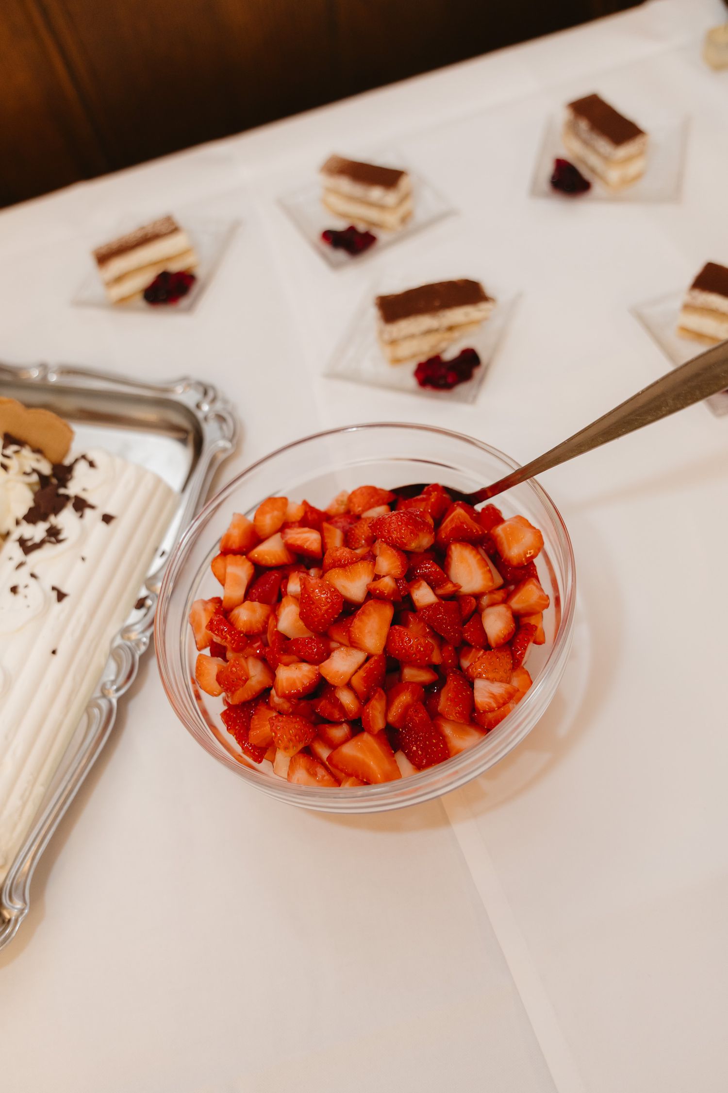 Fresh chopped strawberries in a glass bowl with dessert pastries arranged on white tablecloth in background.