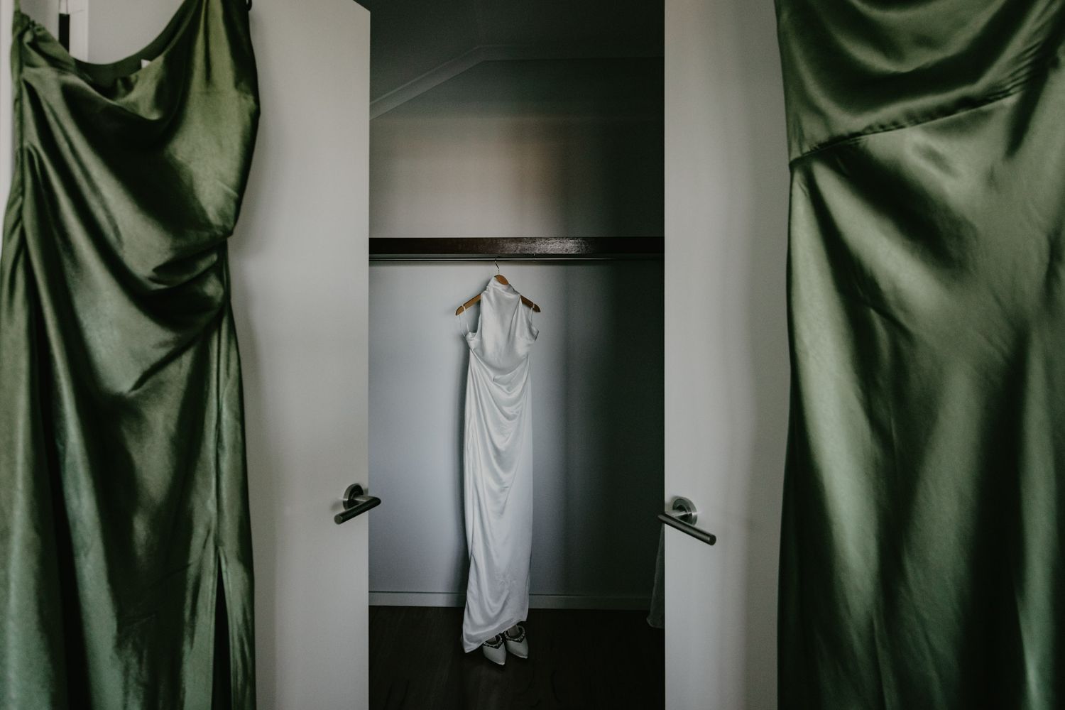 Green bridesmaids dresses hang on doors opening into cupboard with light highlighting brides wedding dress and shoes