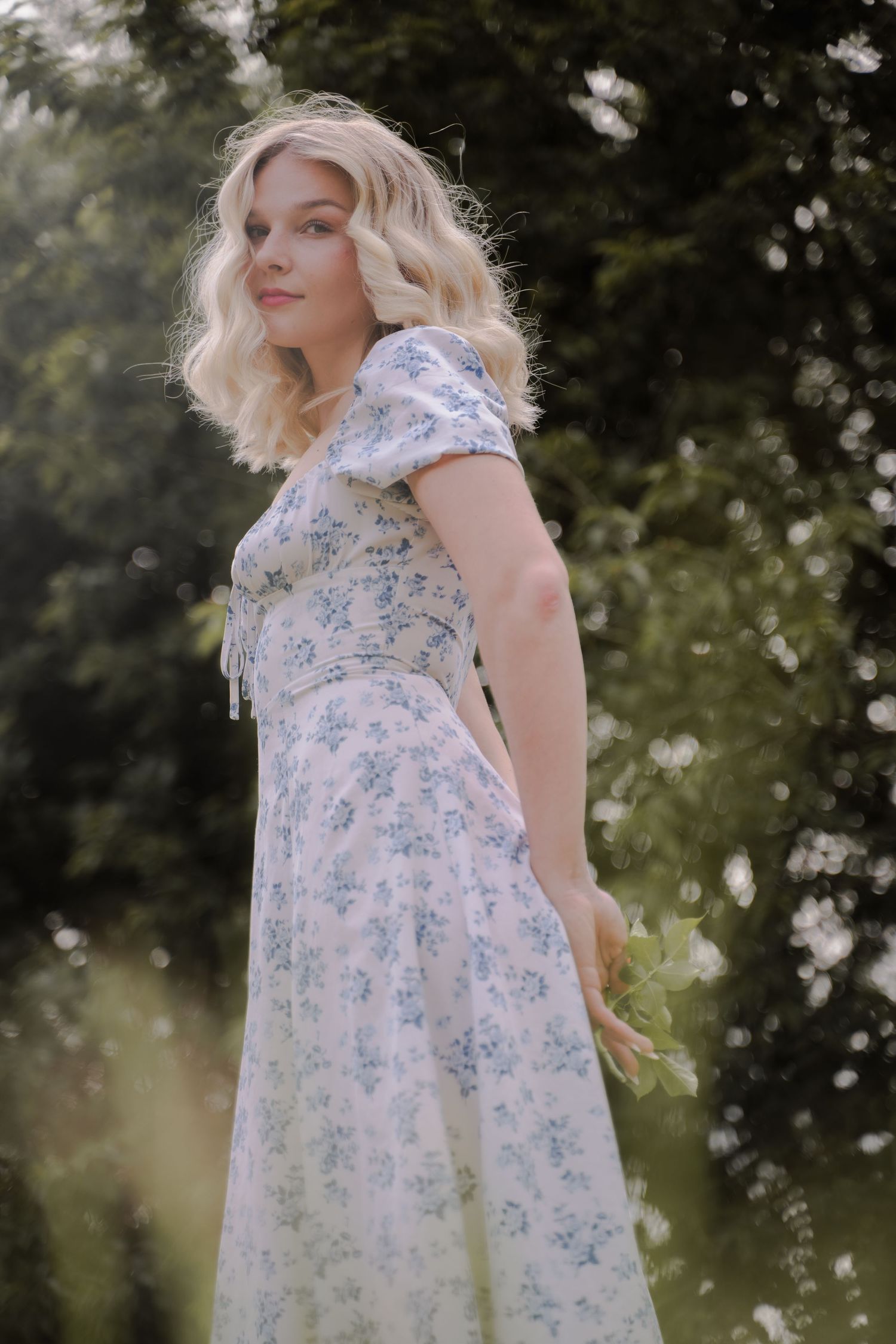 A person in a white and blue floral dress stands in a dreamy outdoor setting with soft bokeh lighting effects.