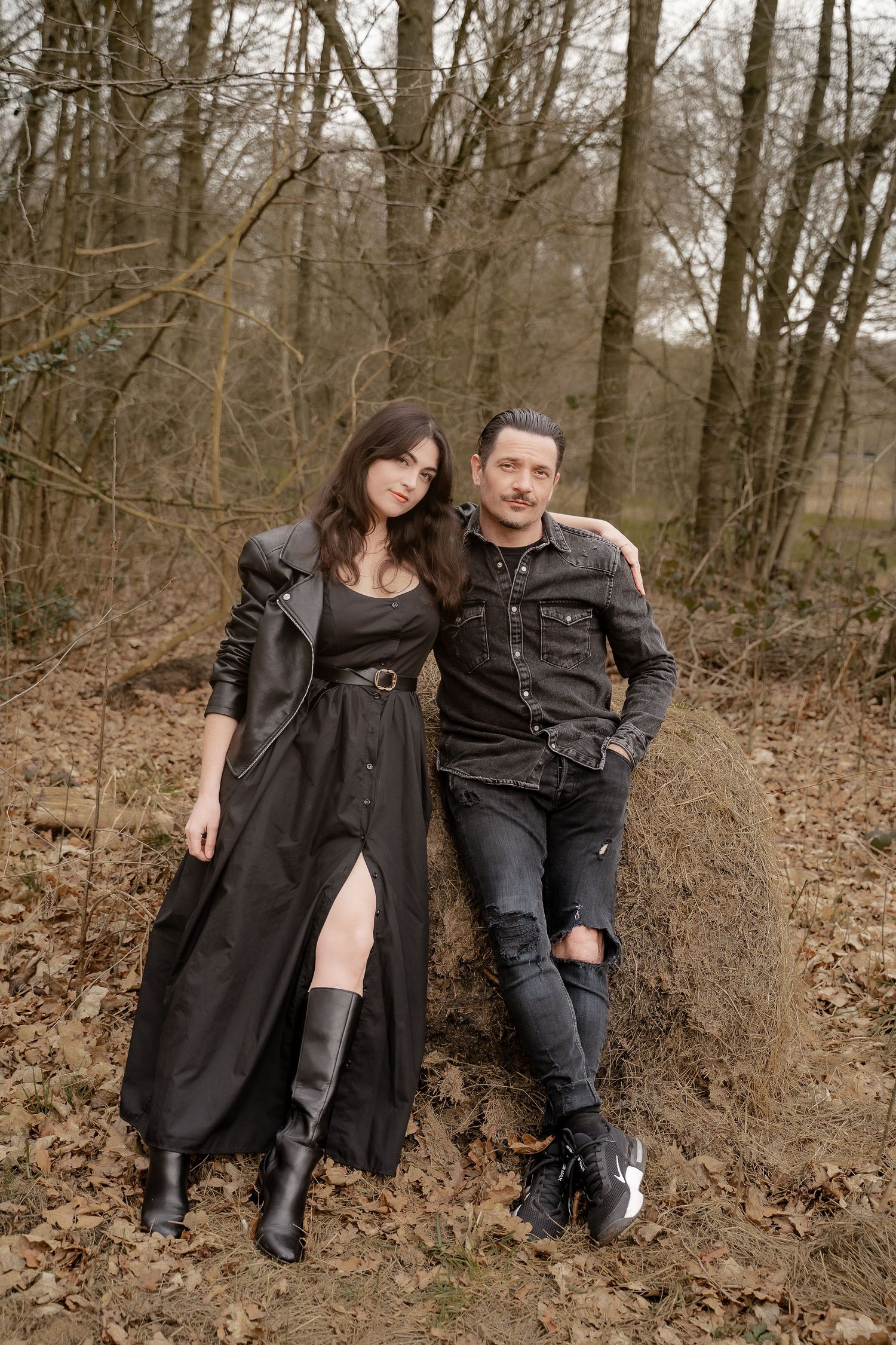 A couple in dark clothing sits together on a fallen tree trunk in a wooded forest setting.