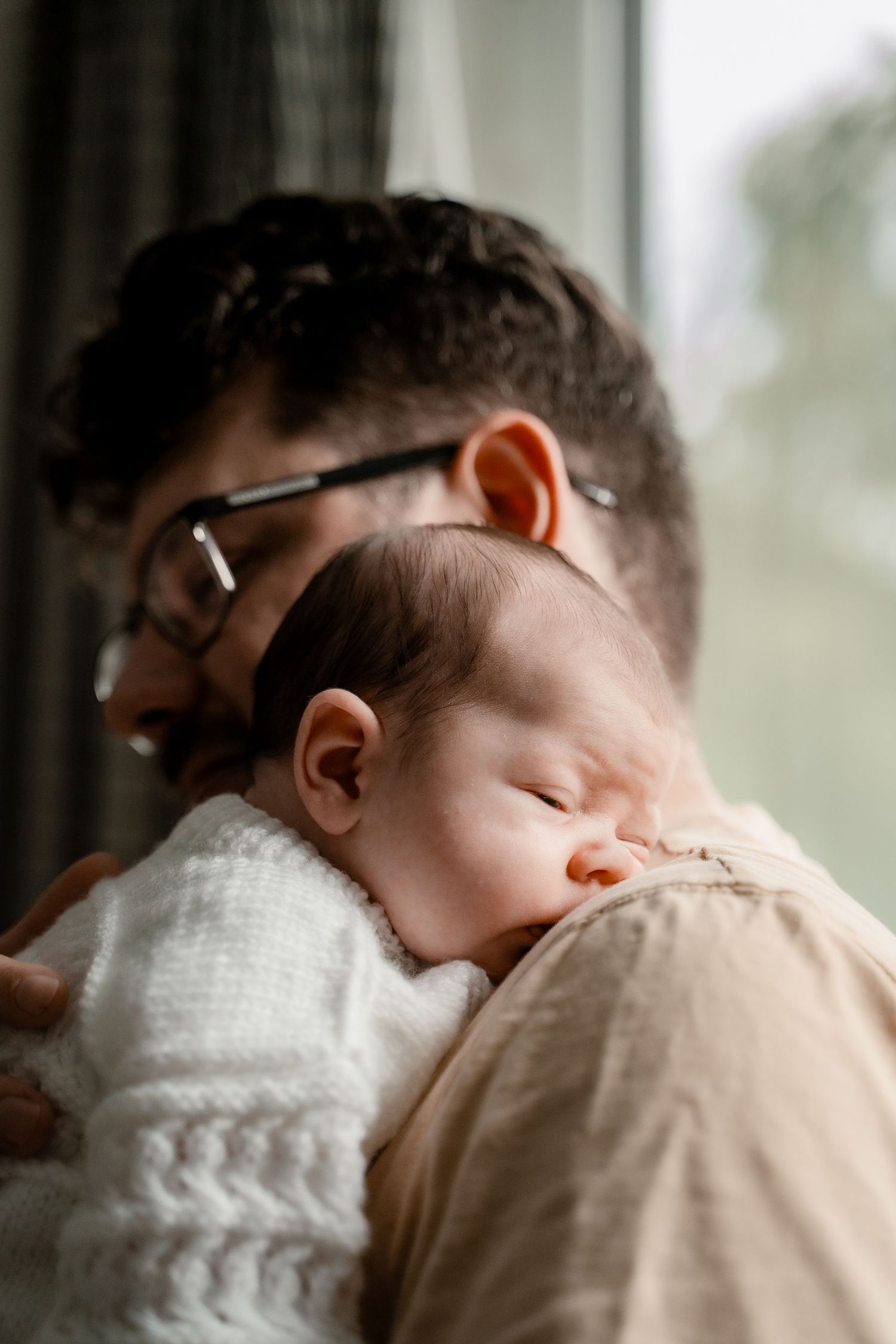 A tender moment shows a sleeping baby in white being held against someone wearing beige.