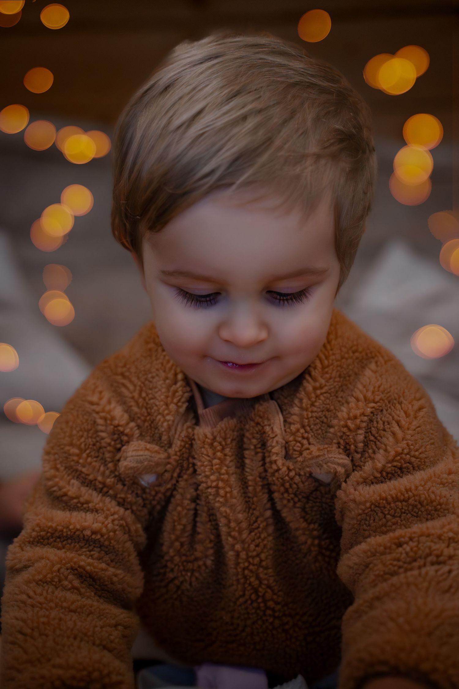 Child in cozy brown teddy bear coat smiling with warm glowing lights in the background.