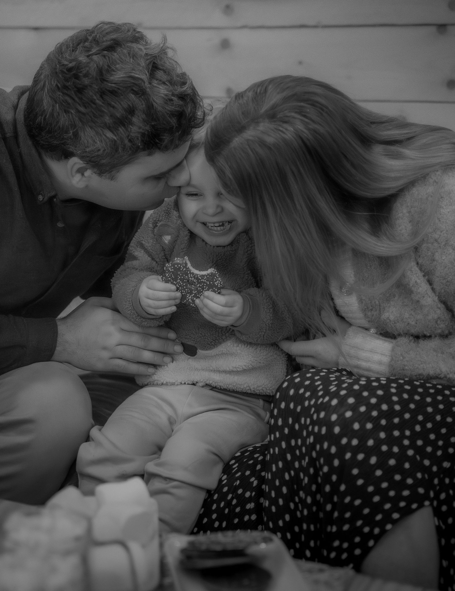 a black and white photo of a mum and dad kissing their child on the cheek In a happy family moment