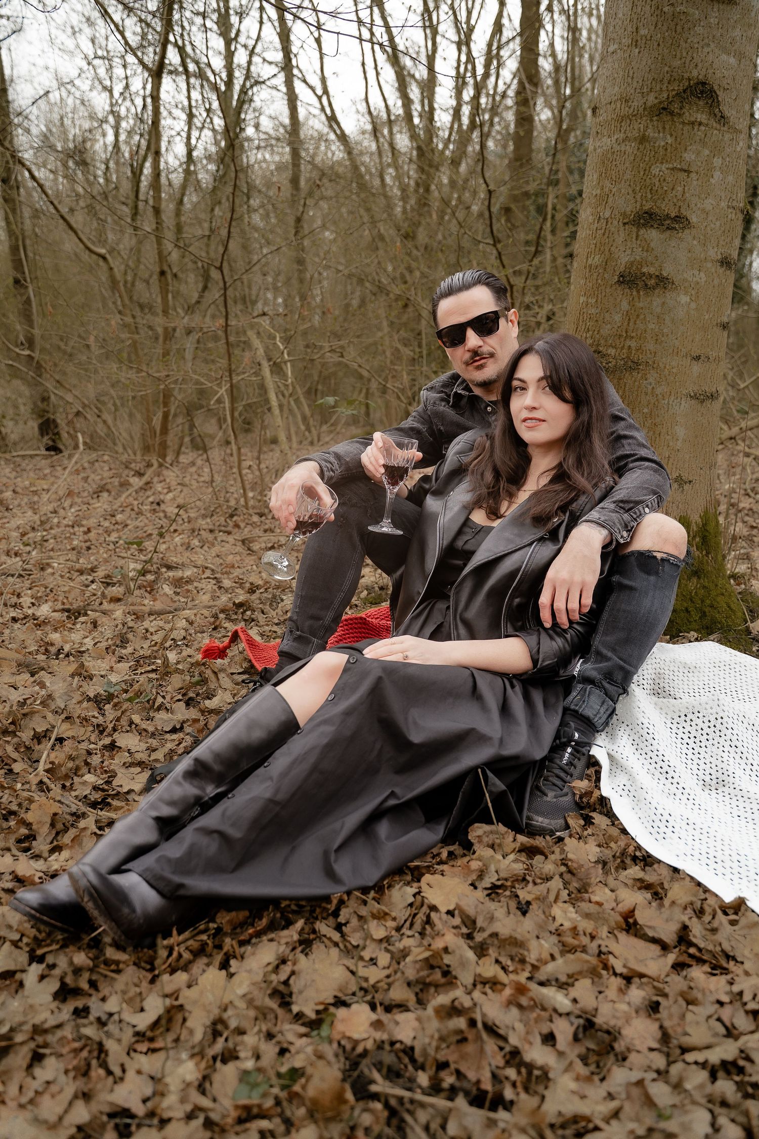 A couple in black leather jackets reclines on autumn leaves in a moody forest photoshoot.