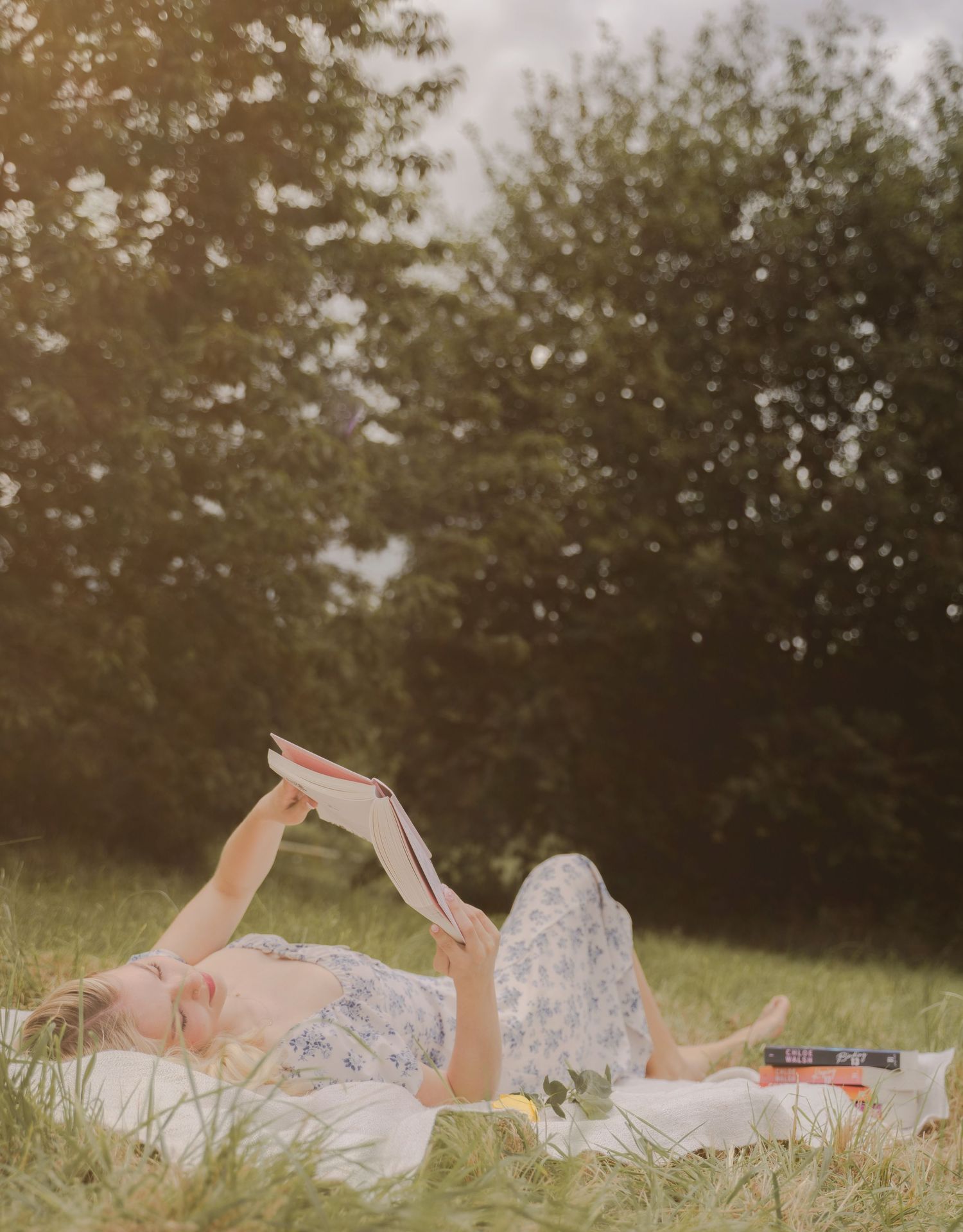 A person in floral dress lies on a grassy field reading a book in a dreamy vintage-style photo series.