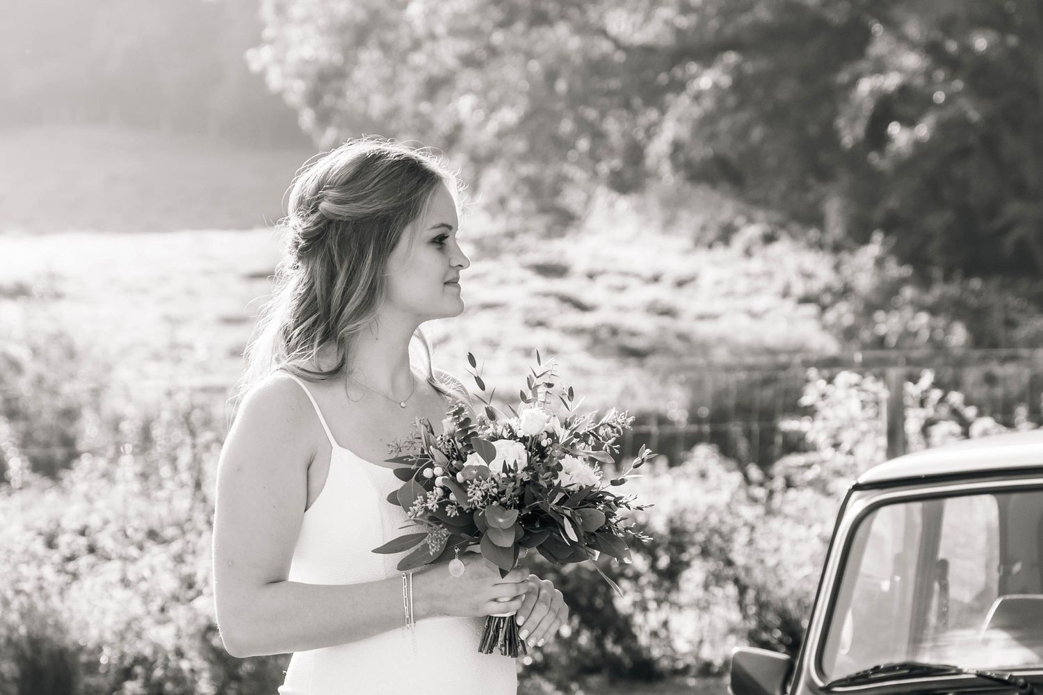 A bride in white holds a lush floral bouquet next to a vintage car in black and white.