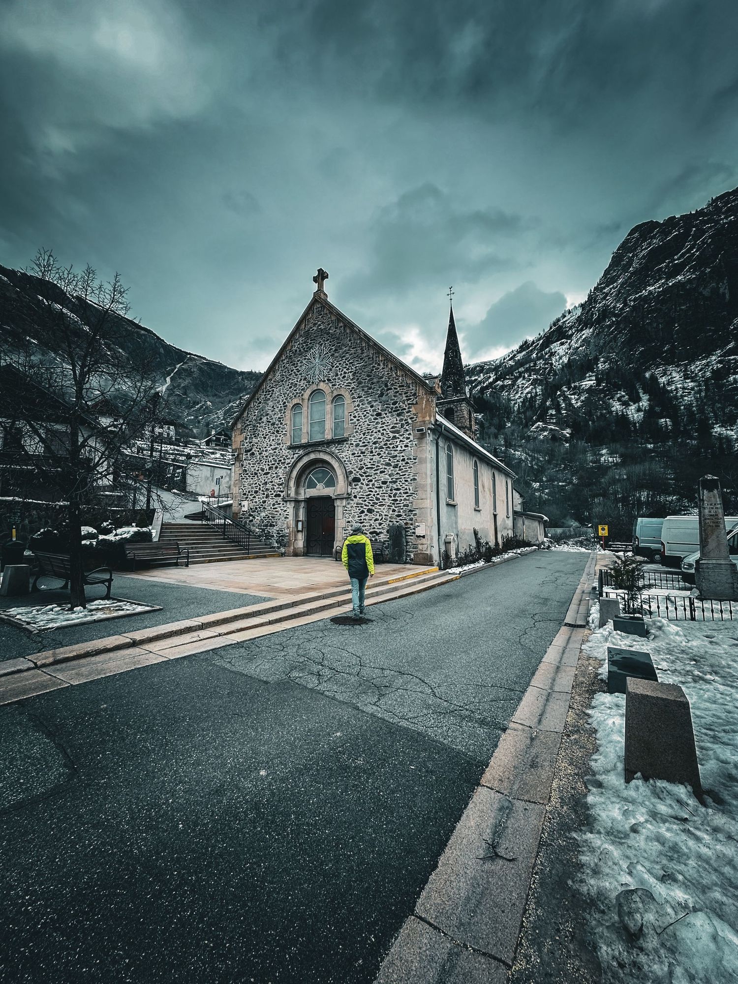 Stone church with spire nestled in a mountain valley with snow-covered peaks in the background.