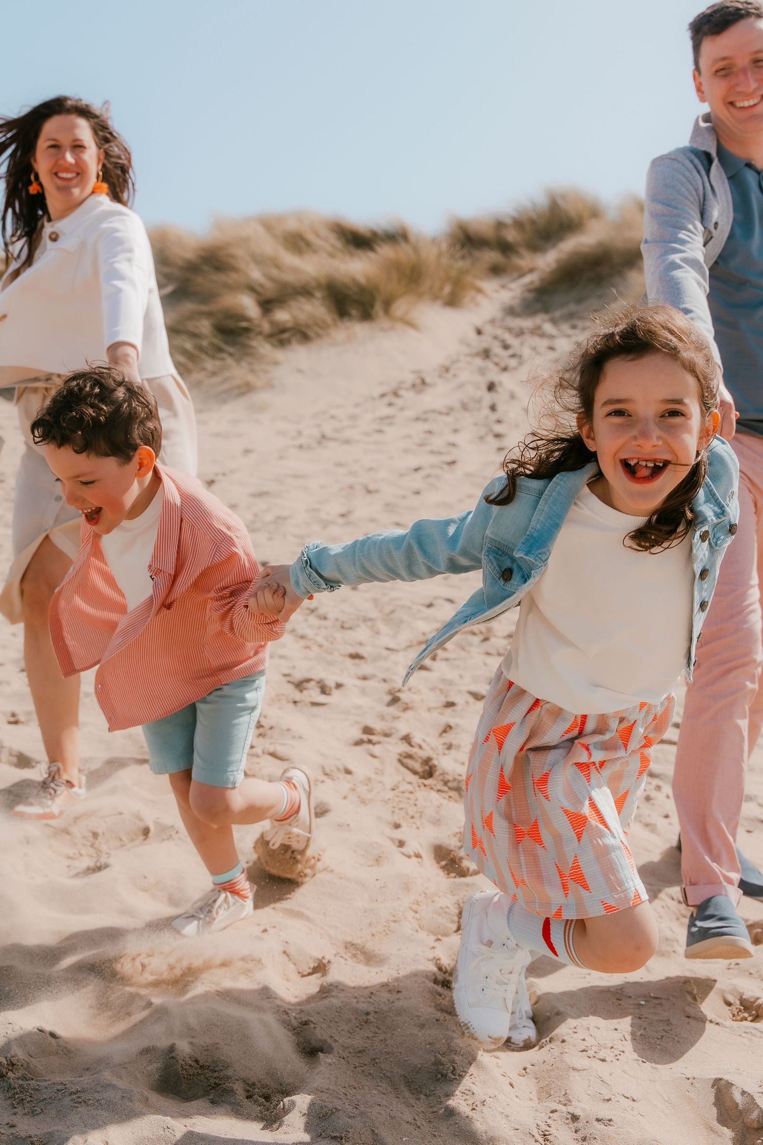 A happy family in coordinating pink and blue outfits playing together on sandy beach dunes at sunset.