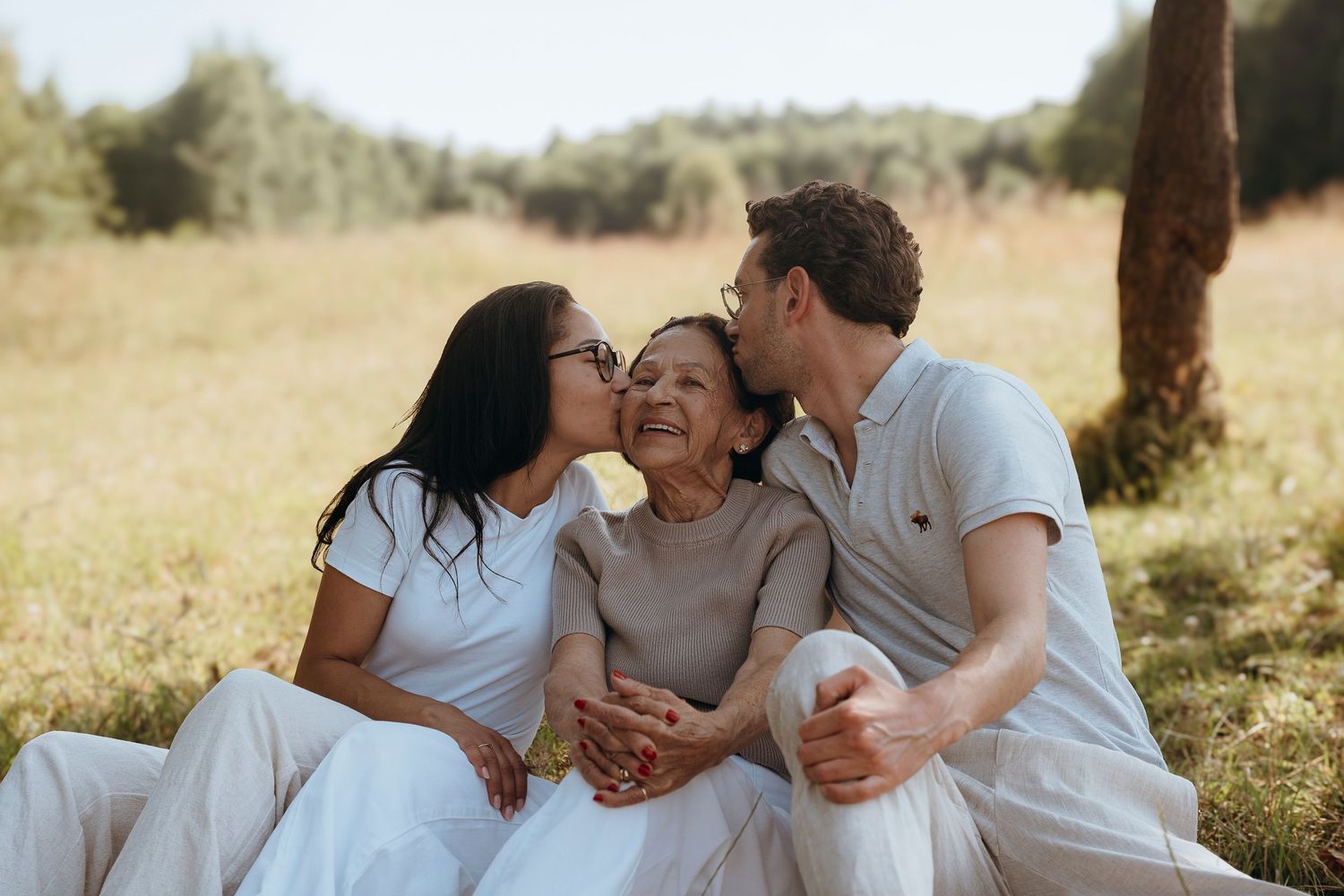 Three people in white clothing share an intimate family moment while sitting together in a sunny field.