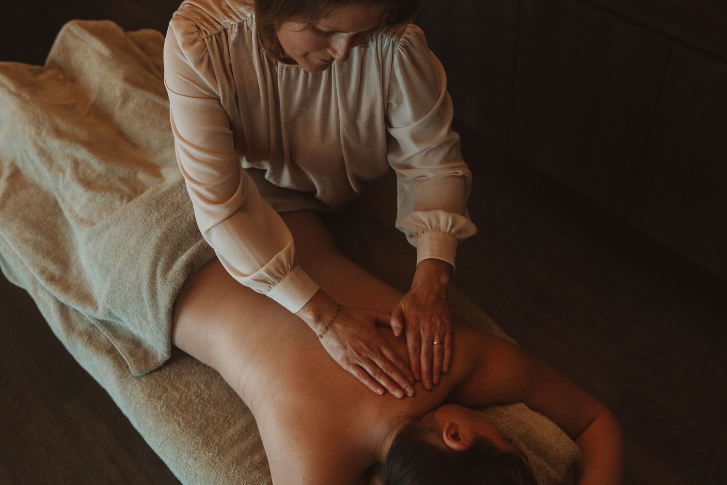 A massage therapist in white attire performs a relaxing back massage treatment on a client in a dimly lit spa room.