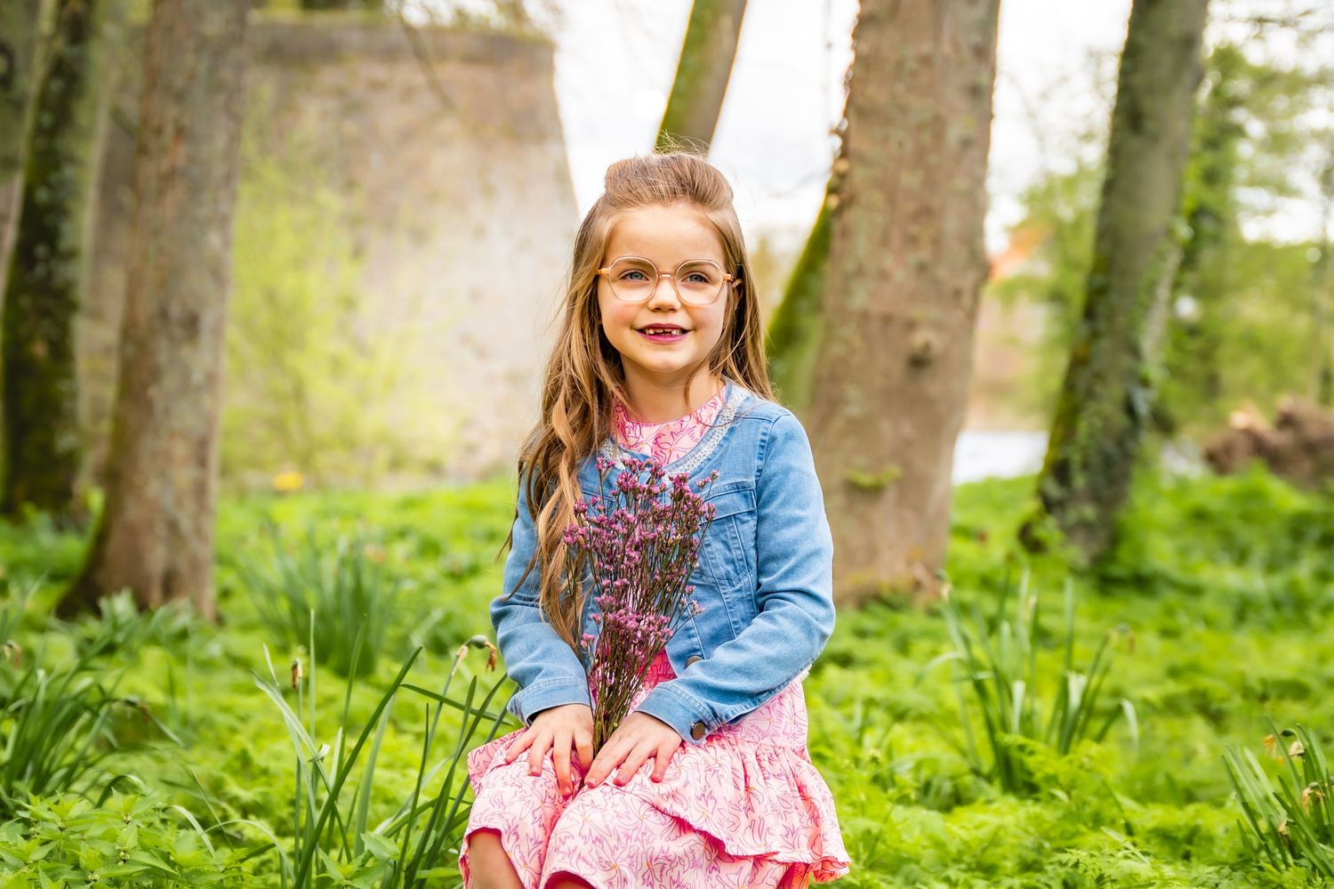Child wearing denim jacket and pink dress sitting in spring forest setting.