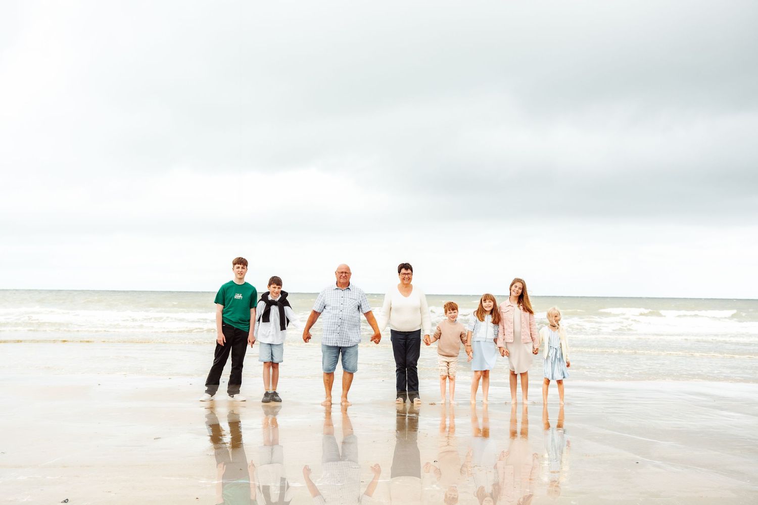 A group walks together along a reflective beach shoreline on an overcast day.