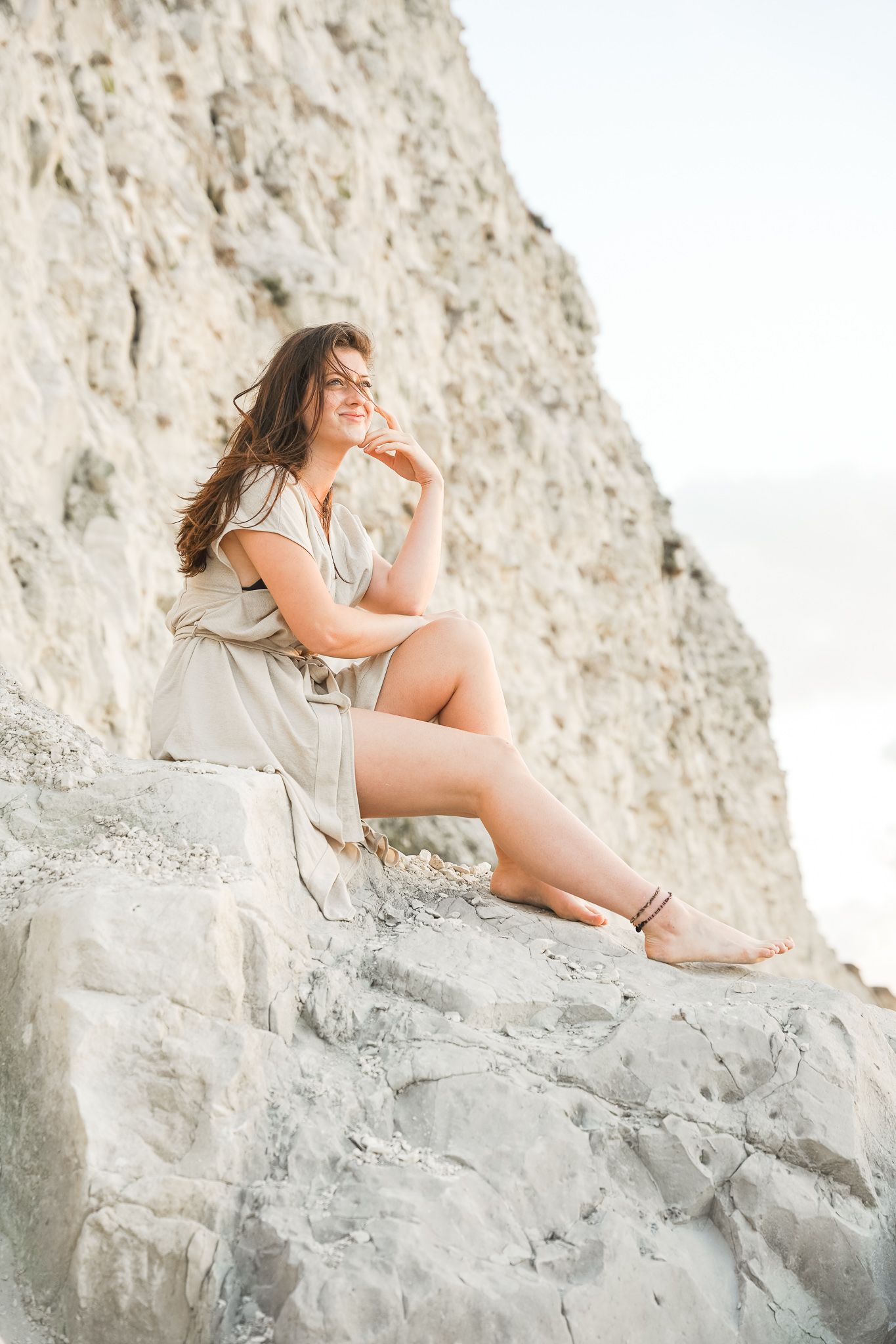 A candid portrait of a person in a beige dress sitting on a white rocky cliff formation during sunset.