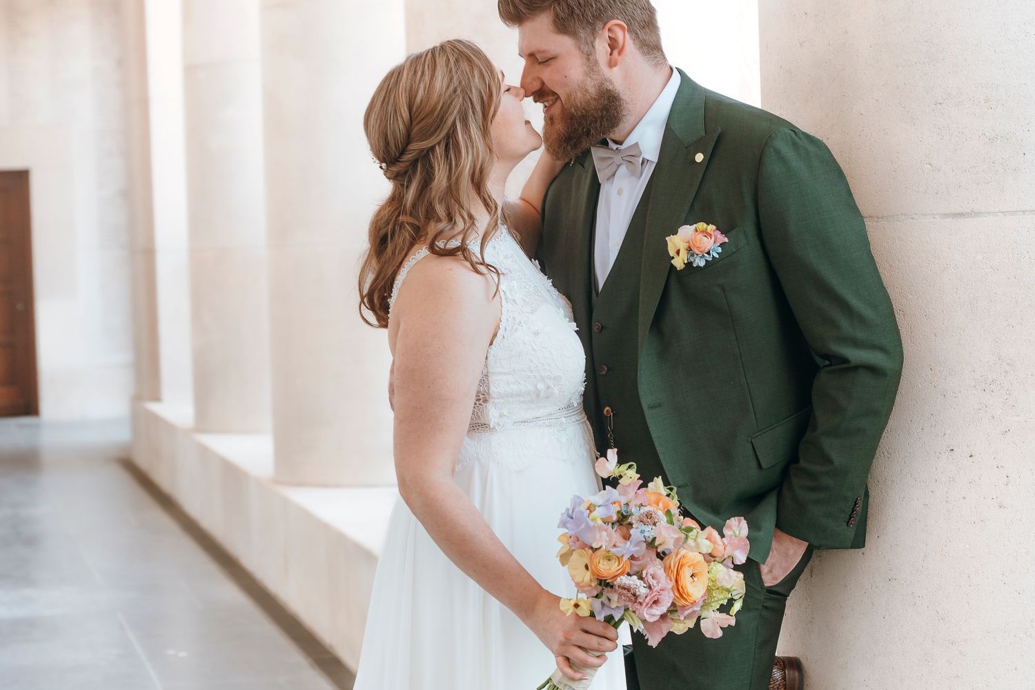 A bride and groom share romantic moments in a bright indoor corridor with white walls and natural lighting.
