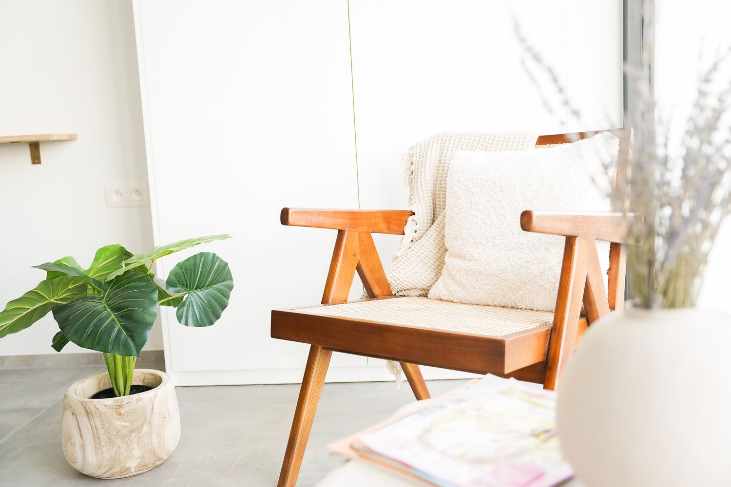 Modern wooden accent chair with cream cushions next to potted plant in minimalist room setting.