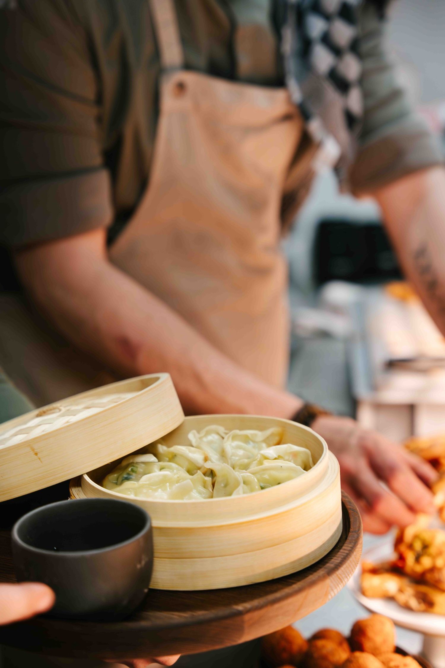 Close-up of bamboo steamer basket with dumplings and black sauce cup on wooden surface.