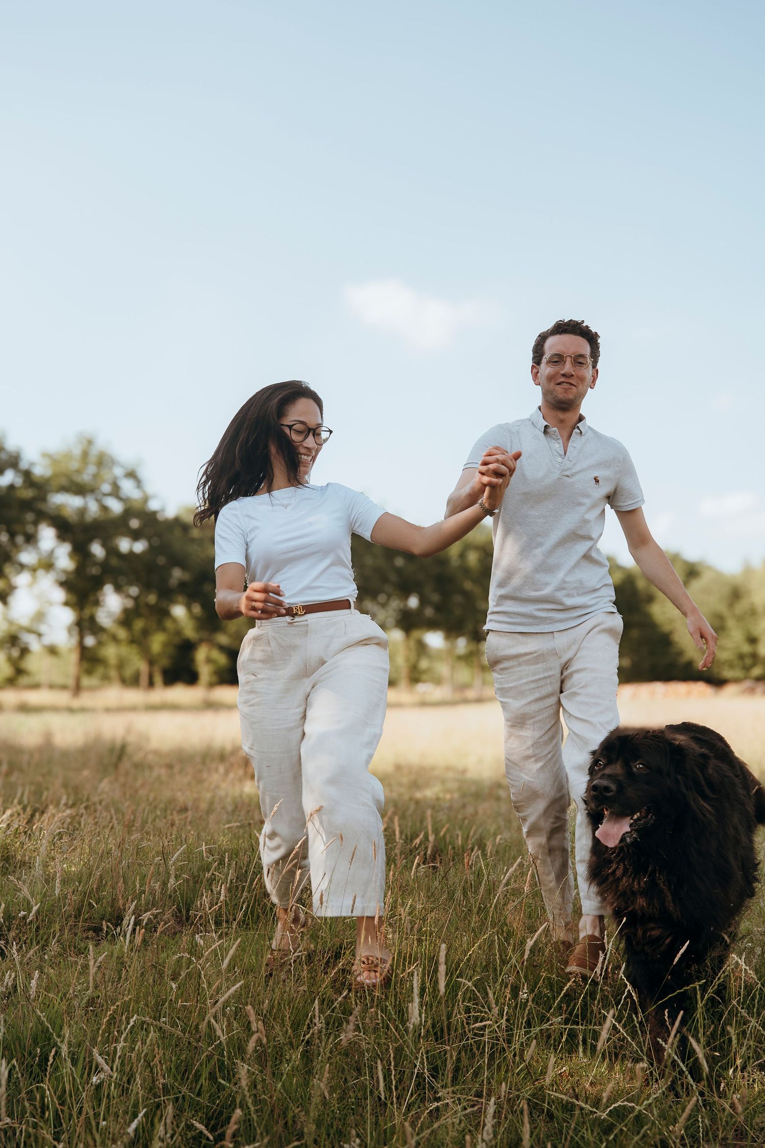A couple in white outfits walks through a grassy field with their large black dog on a sunny afternoon.