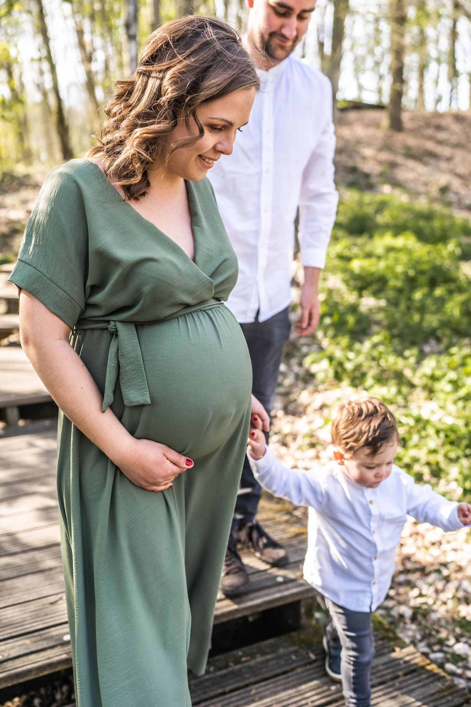 Expecting family stands together on a wooden path in a forest, with mother wearing a green maternity dress.