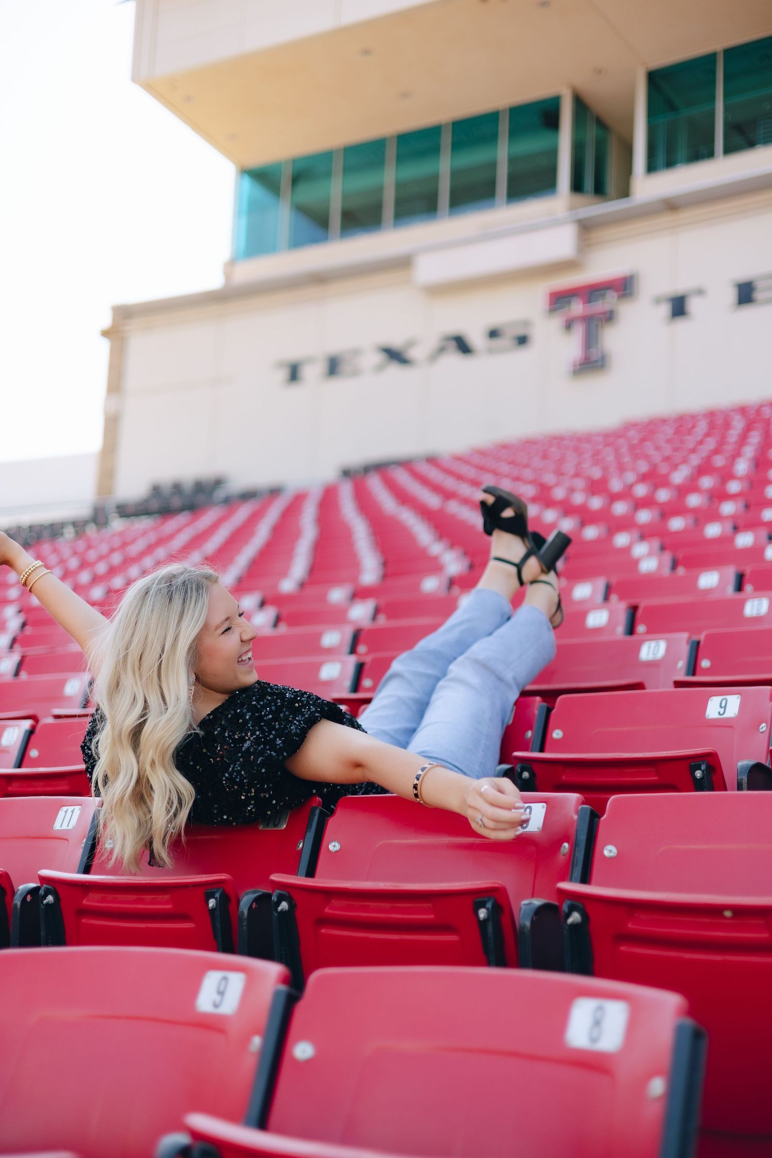 Relaxed pose in light blue jeans at Texas Tech stadium, reclining across empty red stadium seats beneath university signage.