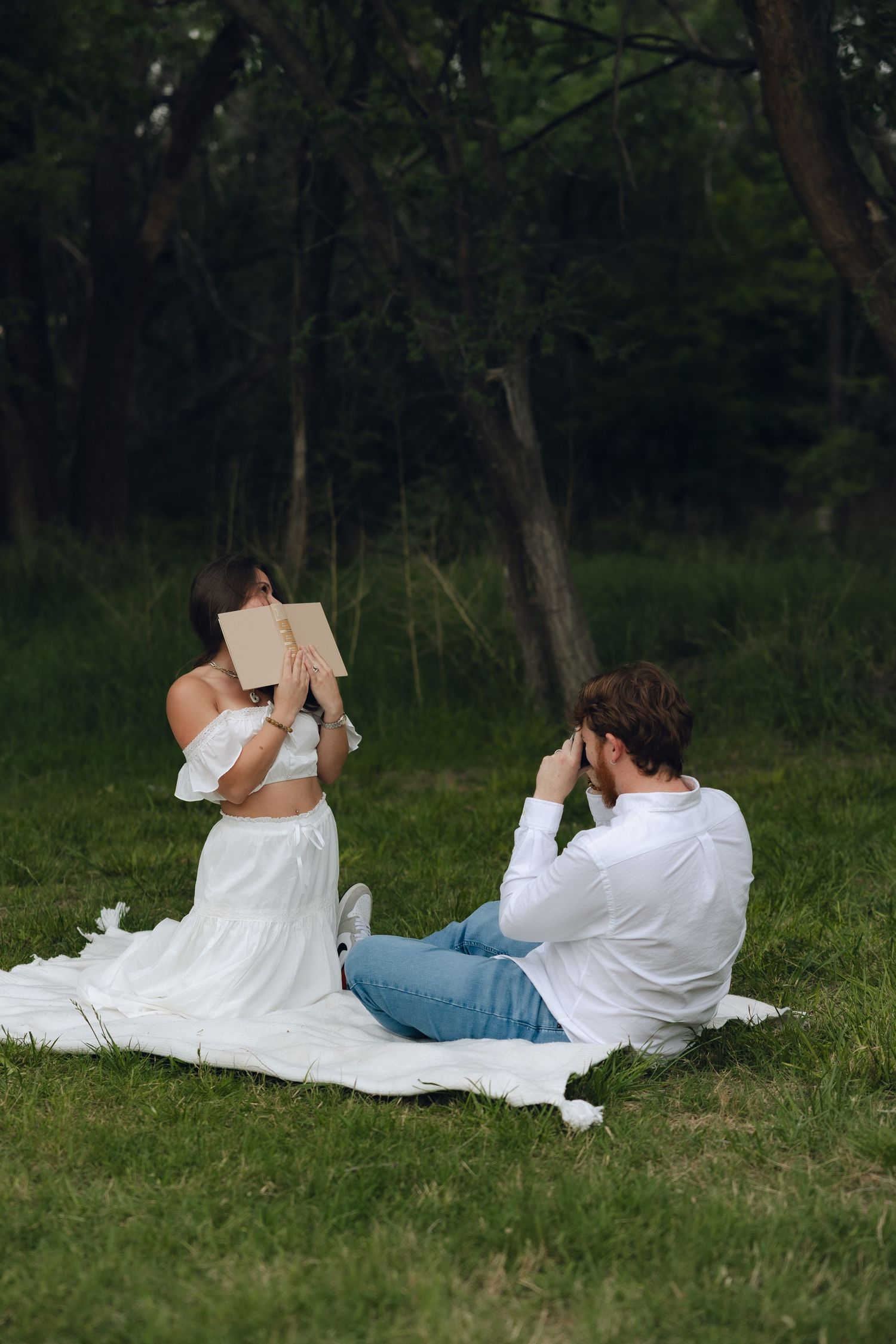 Two people sit on a white blanket in a grassy area reading outside in a wooded setting.