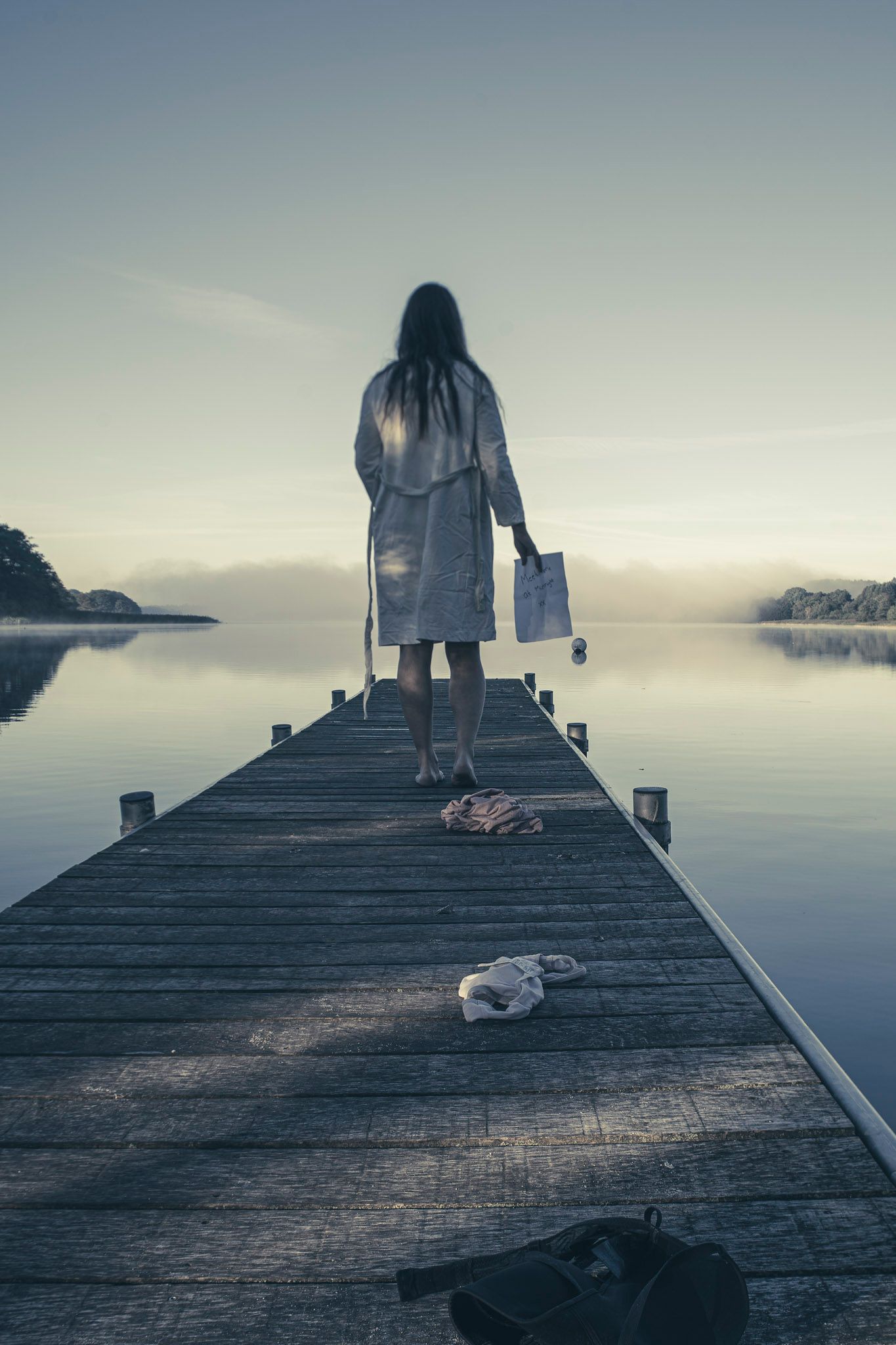 Fotografi af kvinde i badekåbe der står med et brev i hånden på en badebro. Der er tåge i det fjerne