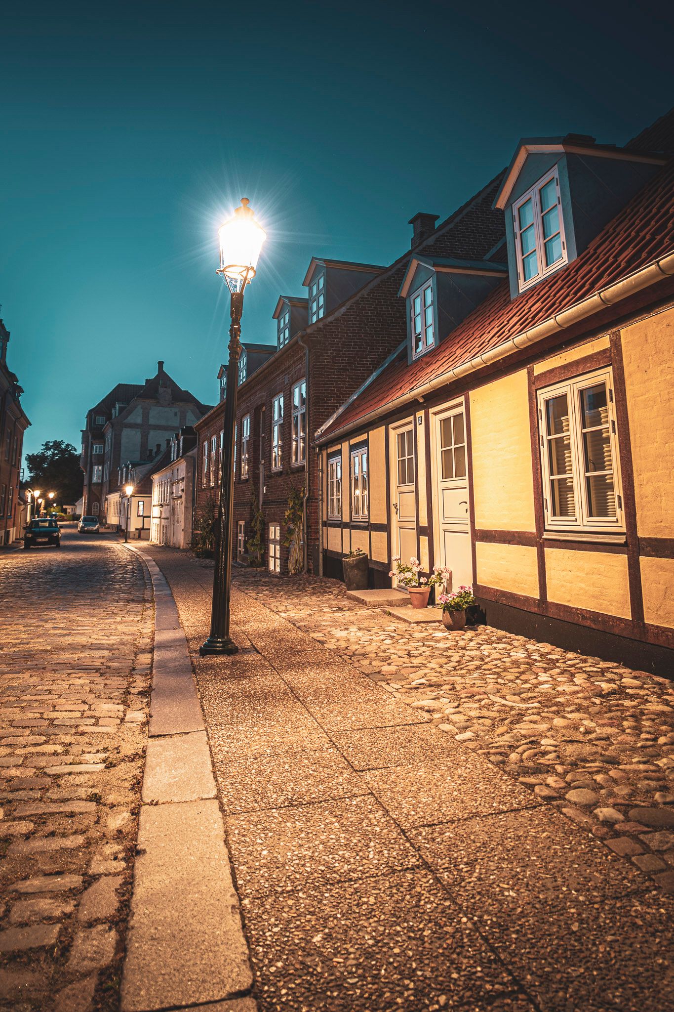 Charming cobblestone street at night with glowing lamp post and historic half-timbered buildings under a teal sky. Sct. Mogensgade in Viborg