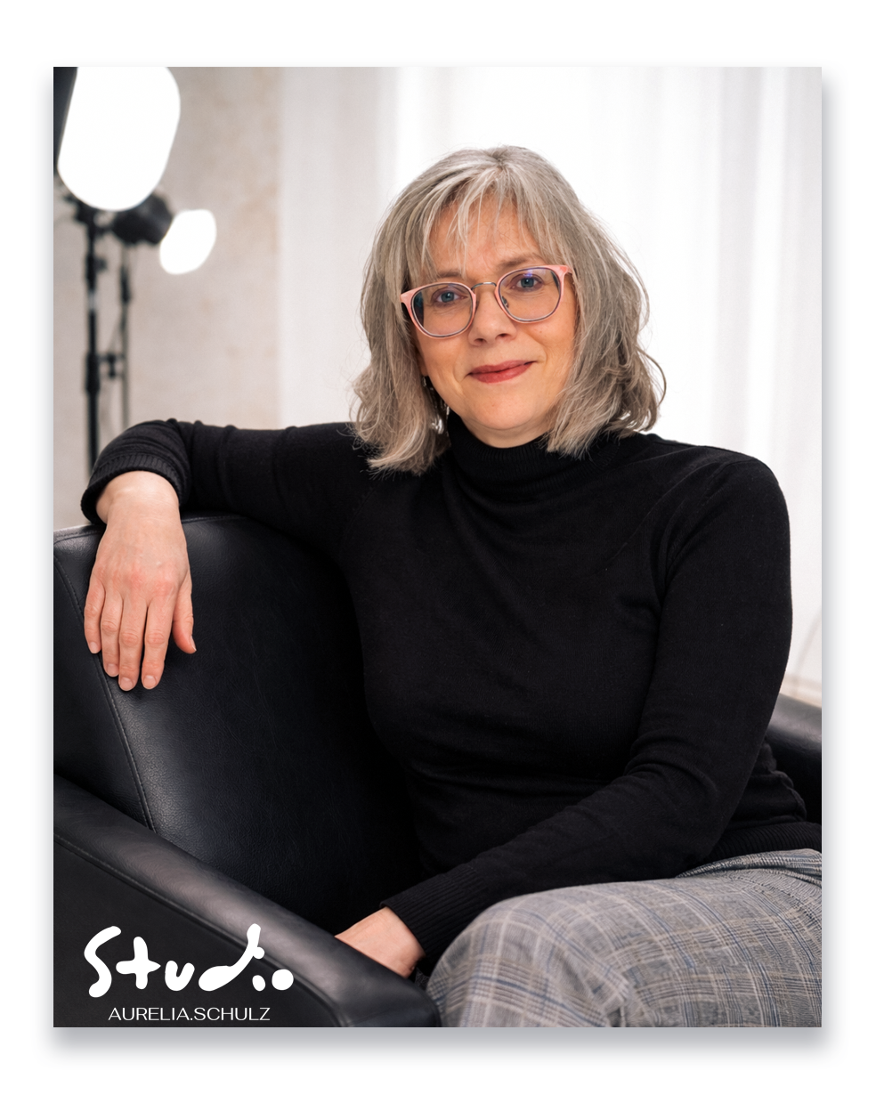A woman with gray hair and glasses sits relaxed in a black leather chair, wearing a black turtleneck, in a Studio Aurelia Schulz portrait.