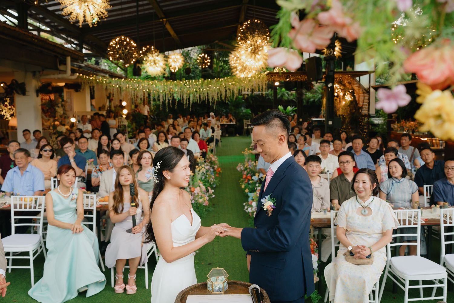 Wedding guests dancing and celebrating on an illuminated outdoor dance floor under string lights and greenery decorations.