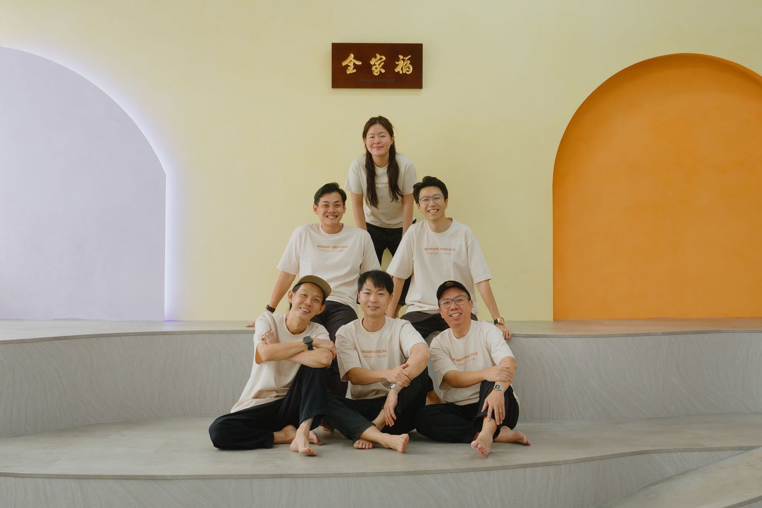 Six people posing in a pyramid formation against a yellow and white wall with Chinese characters above.