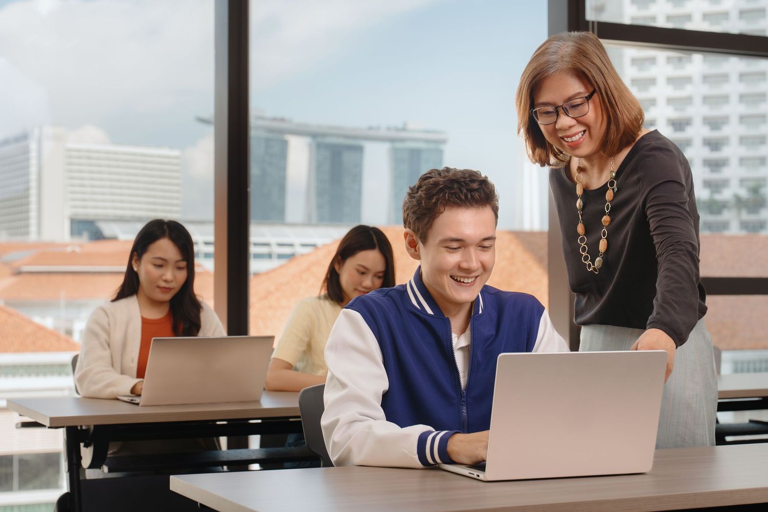 Students collaborate on laptops in a modern classroom, guided by a smiling female teacher in glasses.