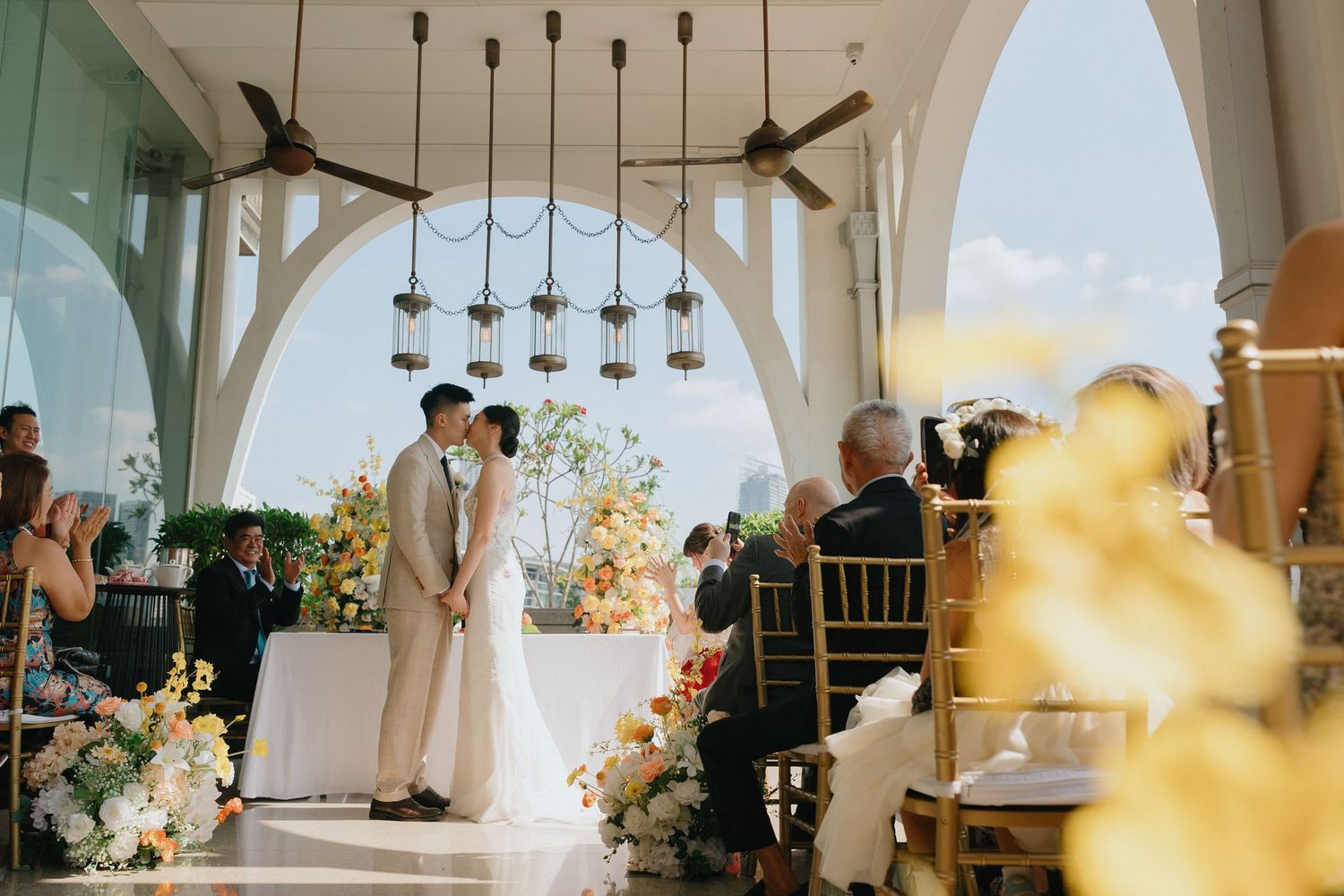 Couple shares a kiss at elegant outdoor wedding ceremony with floral arrangements and hanging lanterns.