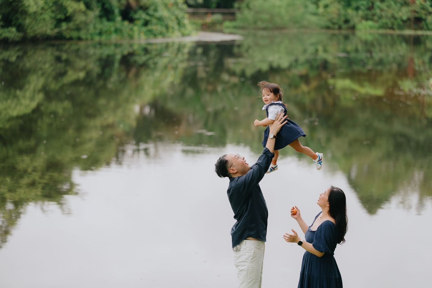 A couple dances playfully by a scenic lake surrounded by lush greenery and trees in a romantic outdoor setting.