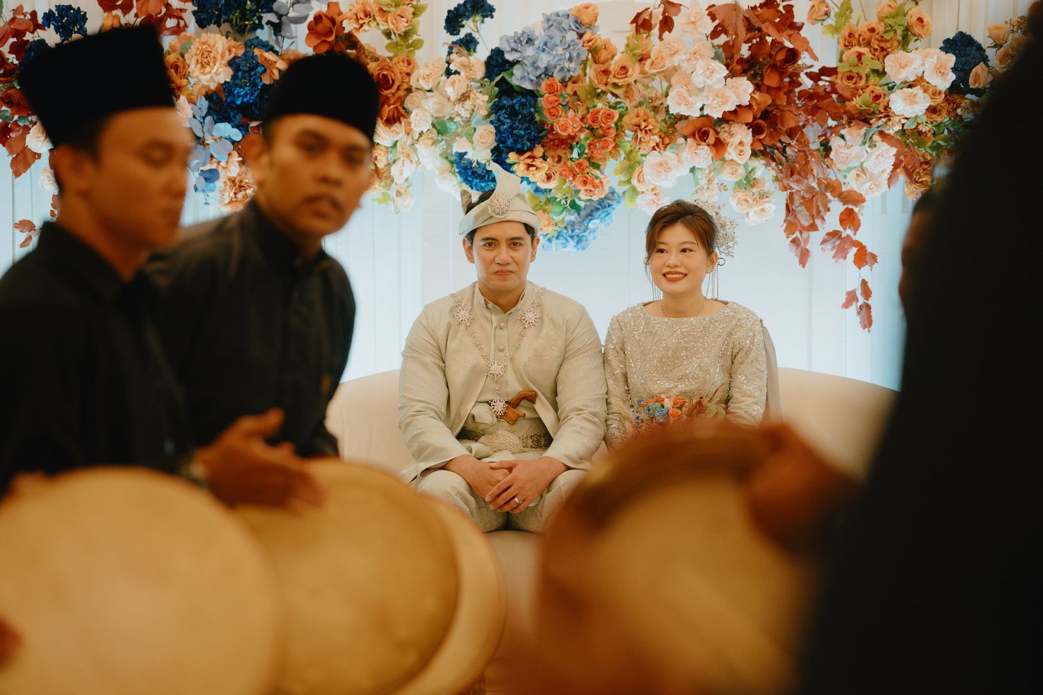 A warm-toned traditional Indonesian wedding ceremony with figures dressed in cultural attire beneath autumn-colored decor.